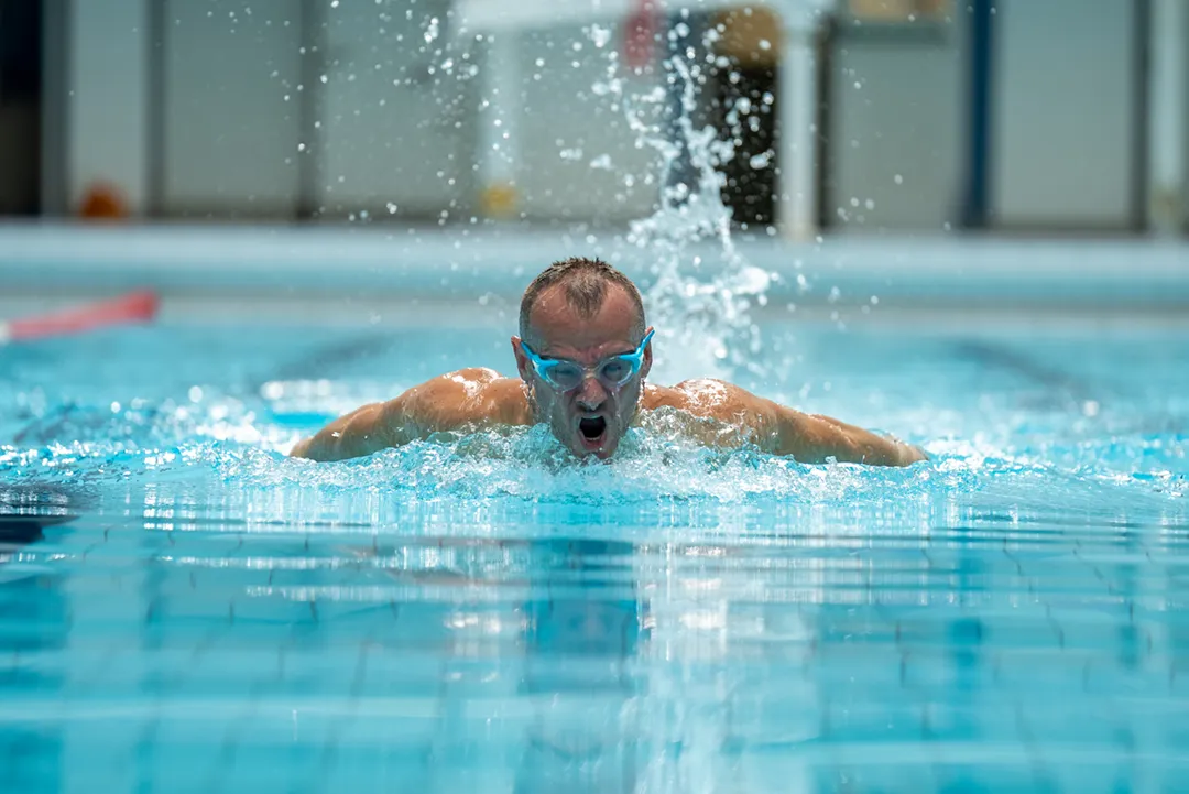 A swimmer wearing blue goggles performs the butterfly stroke in a pool, with water splashing around him.