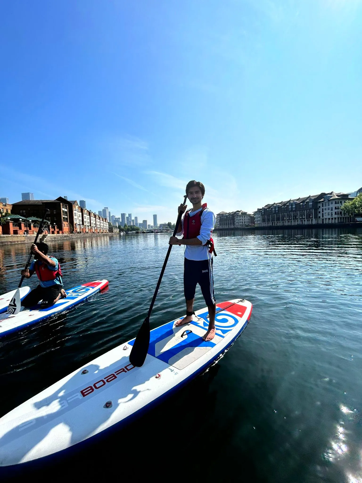 Two paddleboarders on Greenland Docks with blue sky and Canary Wharf view