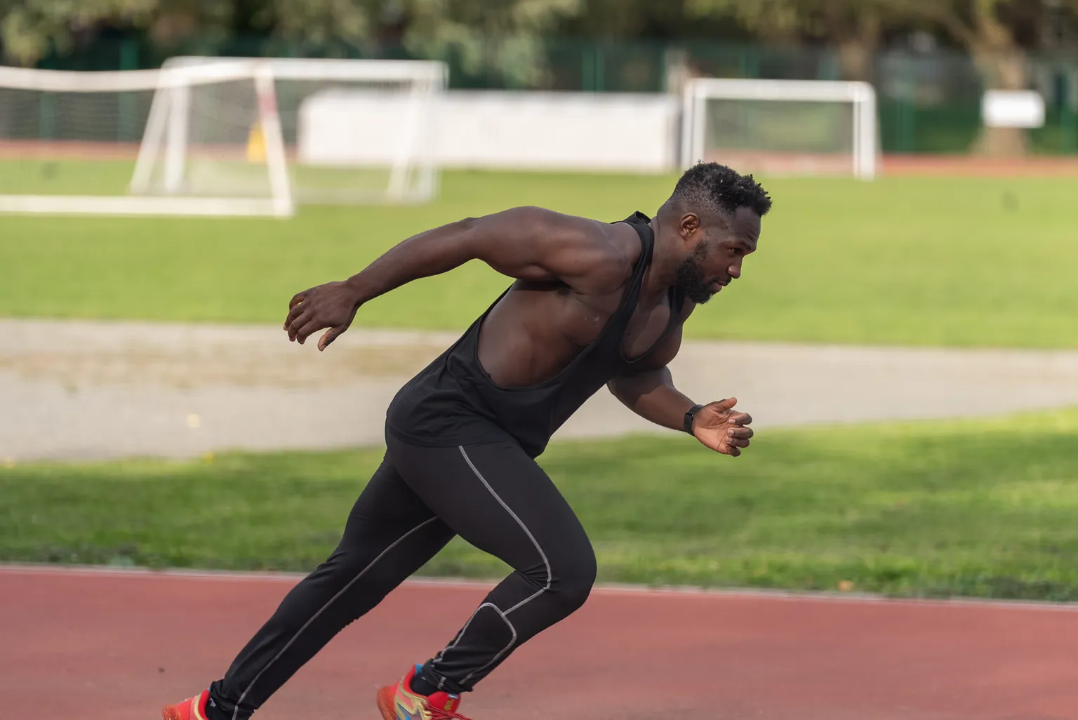 Athlete in a black tank top and leggings sprinting on a track, with green grass and soccer goals in the background.