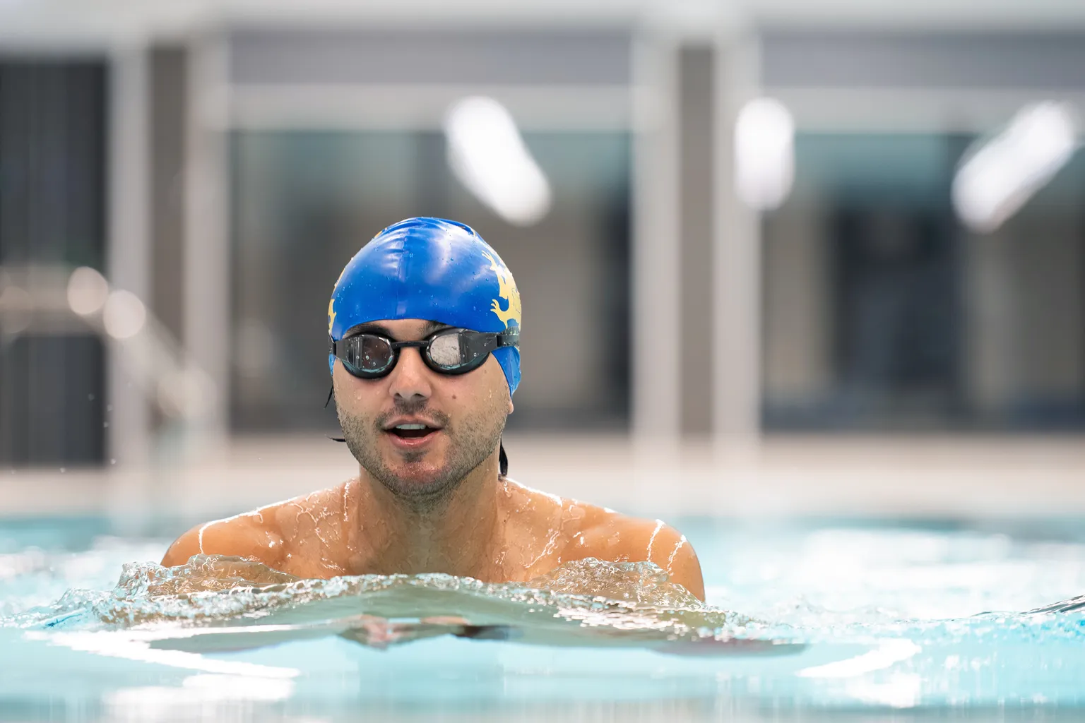 A swimmer in a blue cap and goggles is in a pool mid-stroke, creating ripples in the clear water.