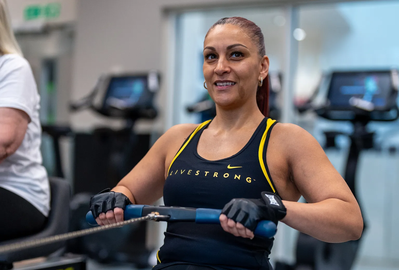Woman exercising on a rowing machine at the gym, wearing a black and yellow tank top and gloves, with a focused expression.