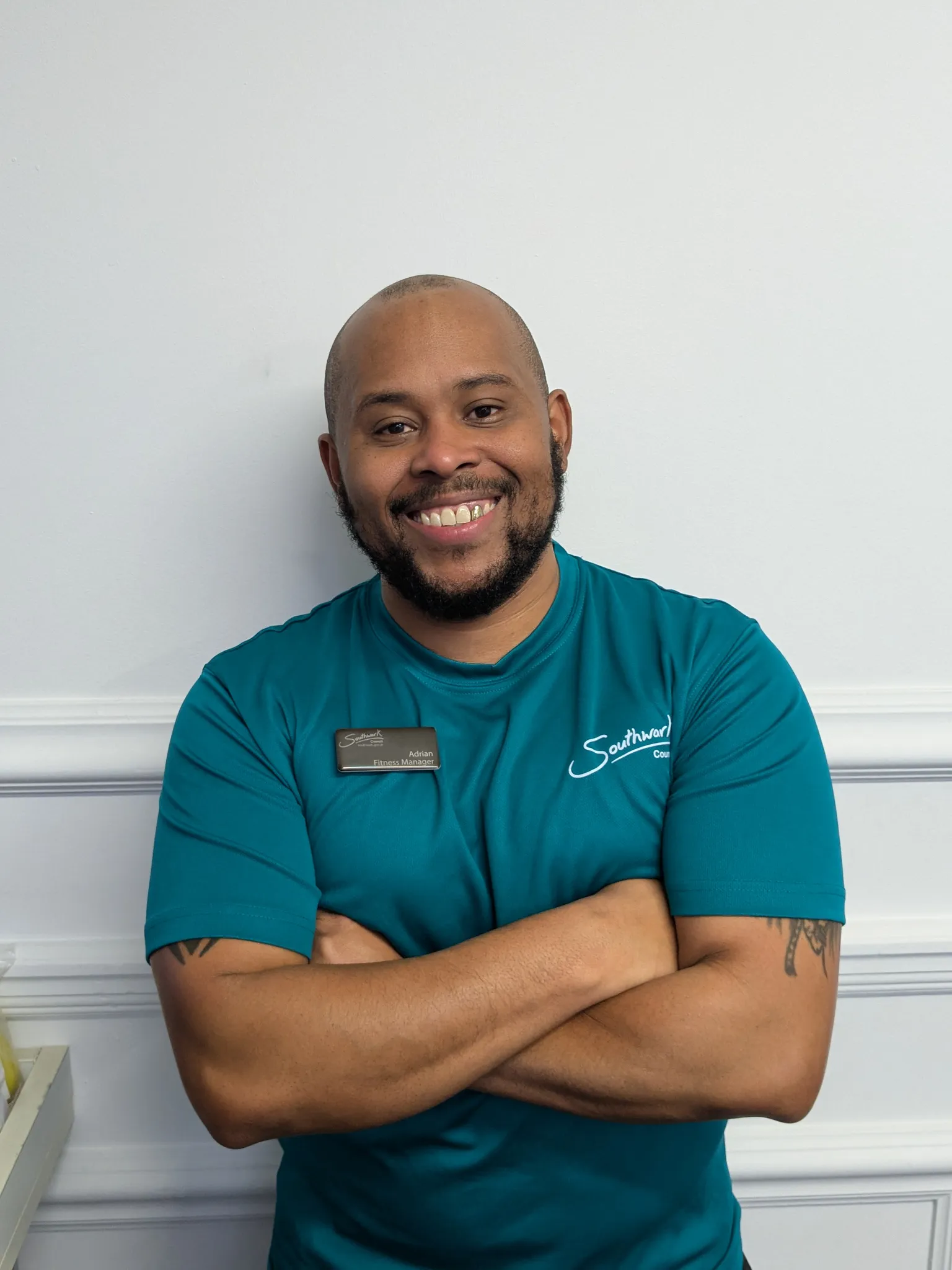 A smiling man with a beard, wearing a teal shirt and a name tag, stands with arms crossed against a white wall.