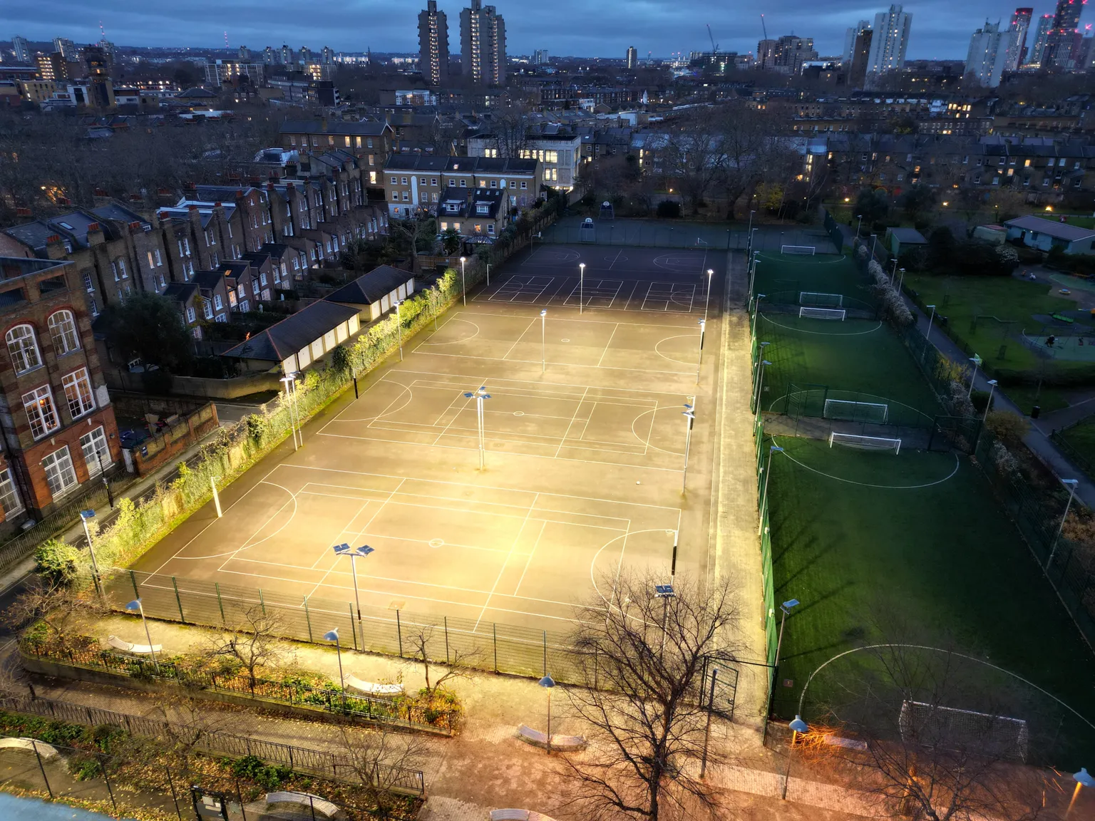 Illuminated sports courts and soccer fields in an urban area at dusk, surrounded by residential buildings and lit streetlights.