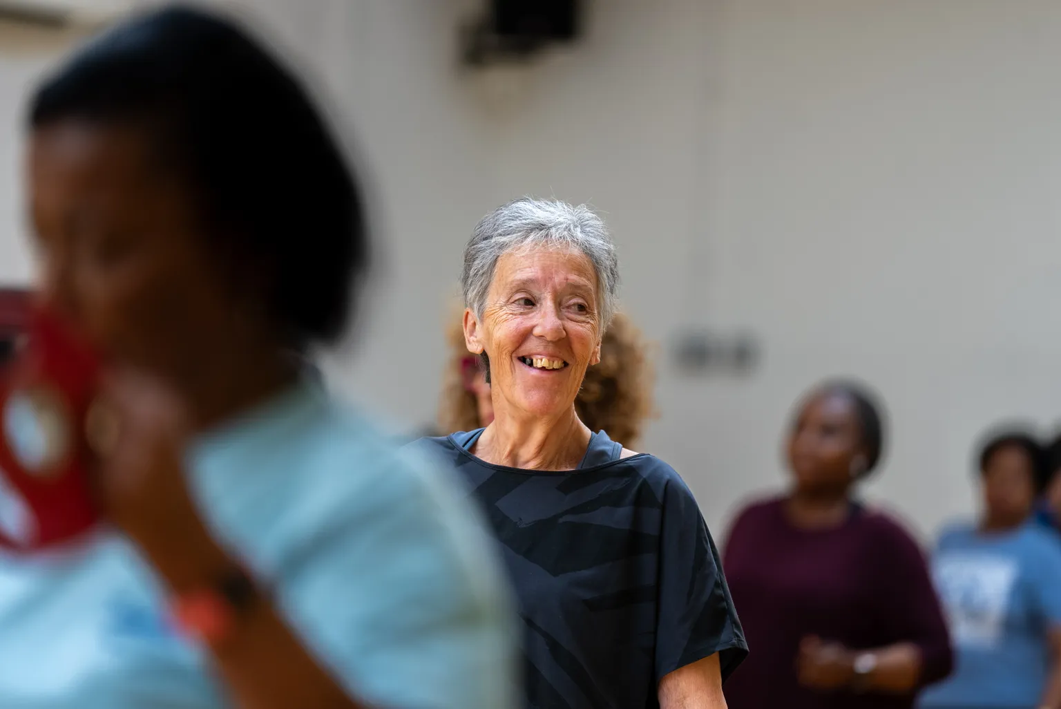 A smiling elderly woman stands in a group setting, appearing happy and engaged, with blurred individuals in the background.