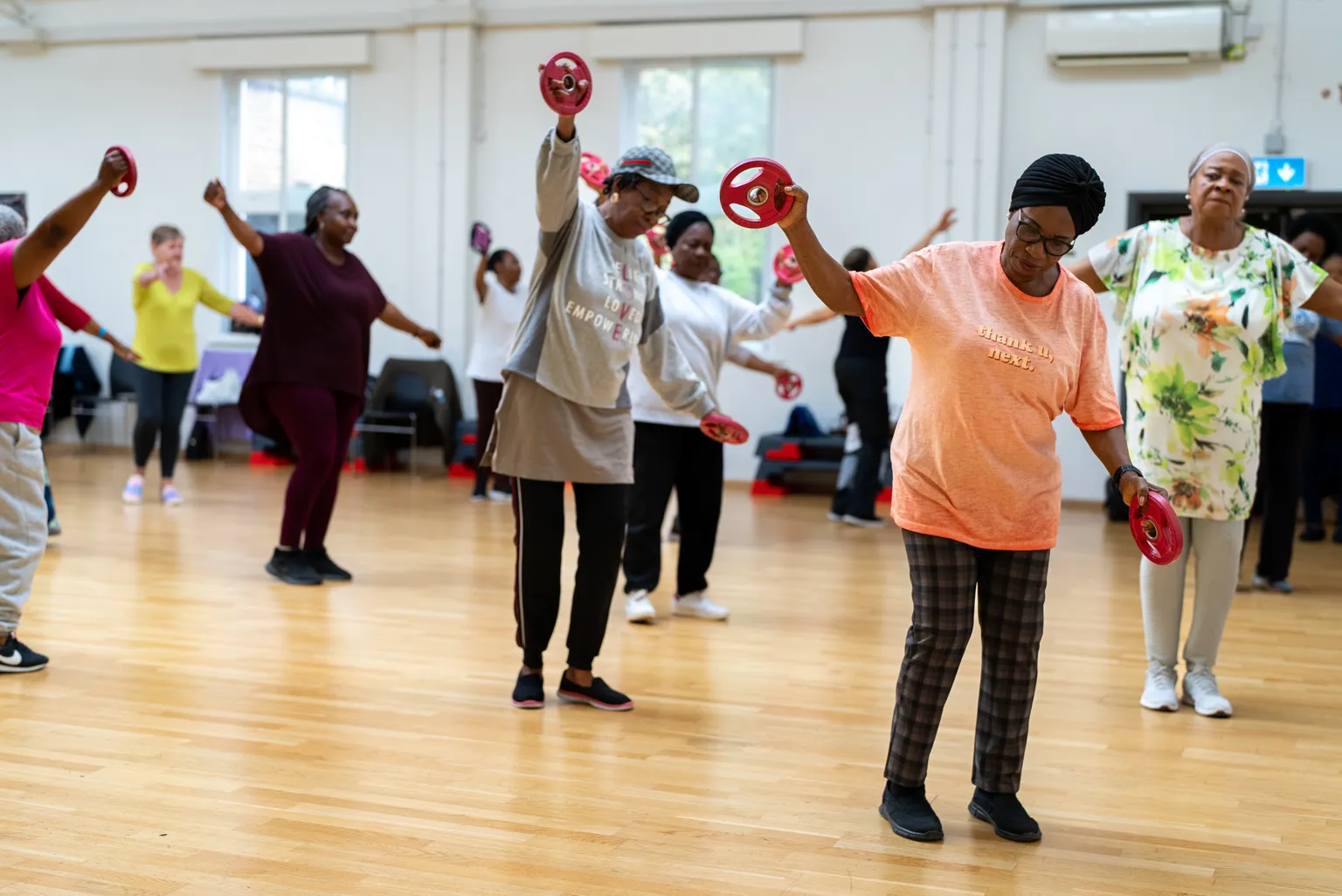 A group of women in a fitness class workout with small red weights, moving energetically in a bright, spacious studio.