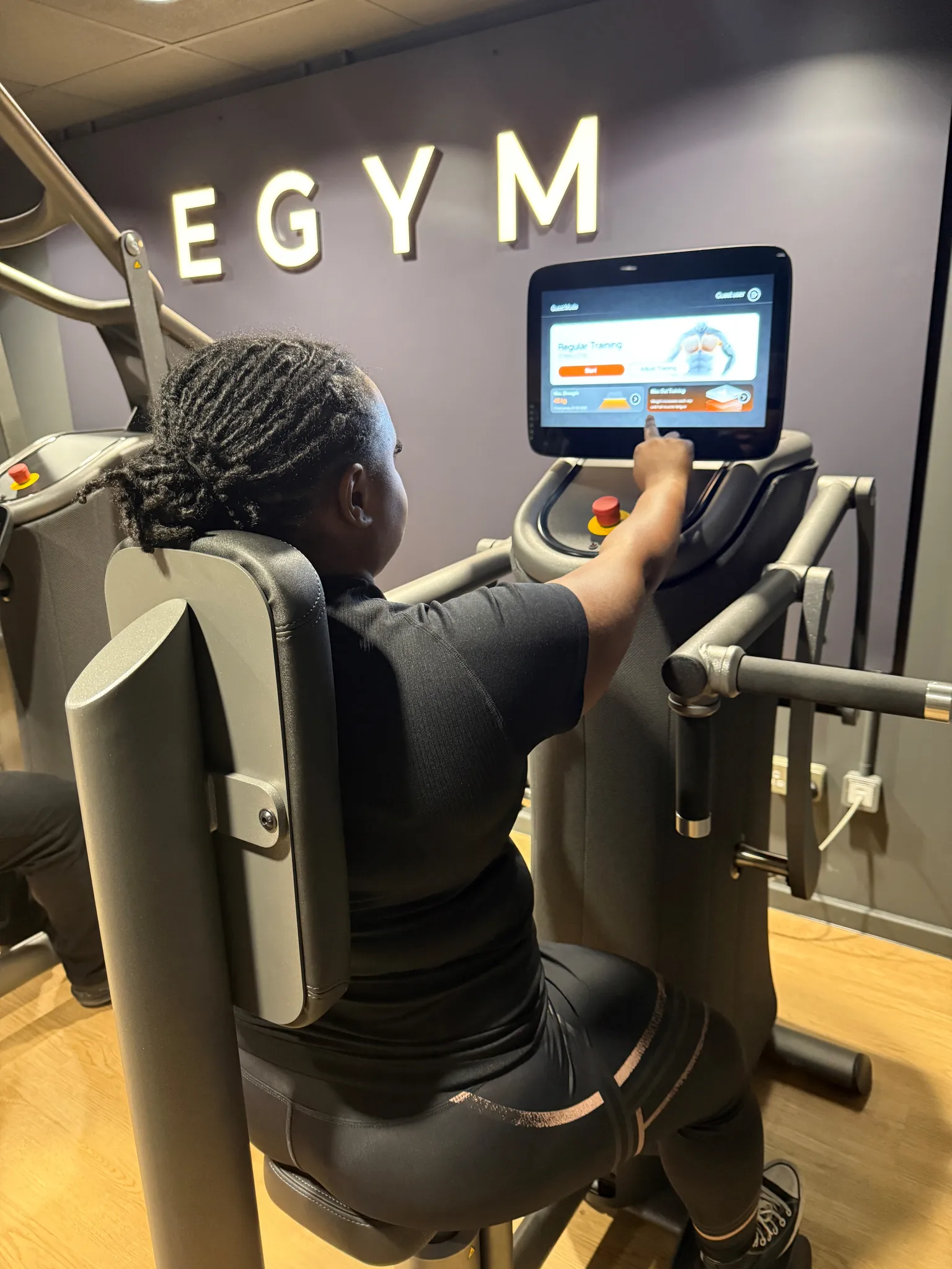 Person using a gym machine with a touchscreen interface labeled "EGYM," selecting a training program in a fitness center.