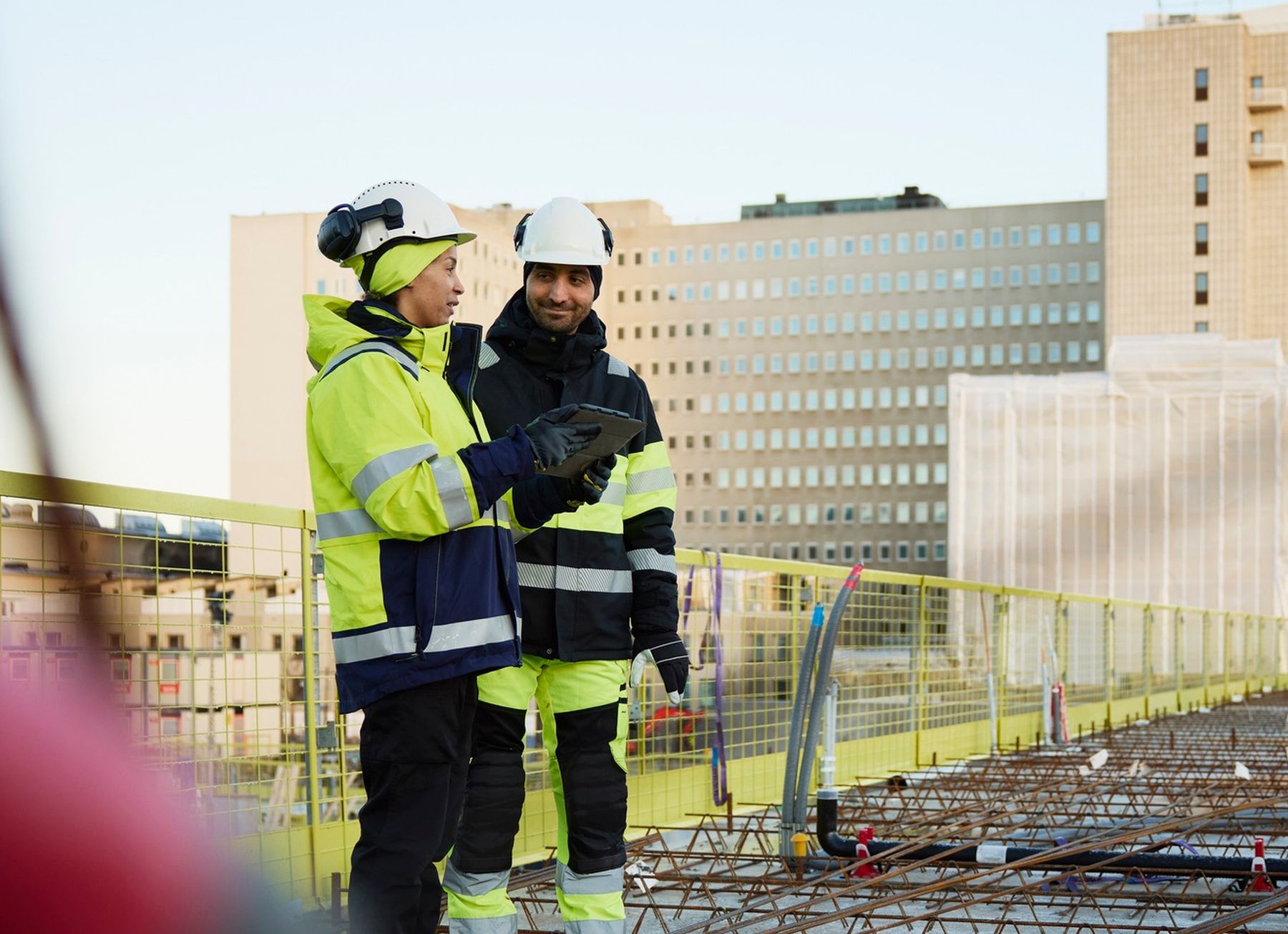 To bygningsarbejdere i reflekstøj og hjelme står på en byggeplads og diskuterer noget på en tablet, mod en bybaggrund.