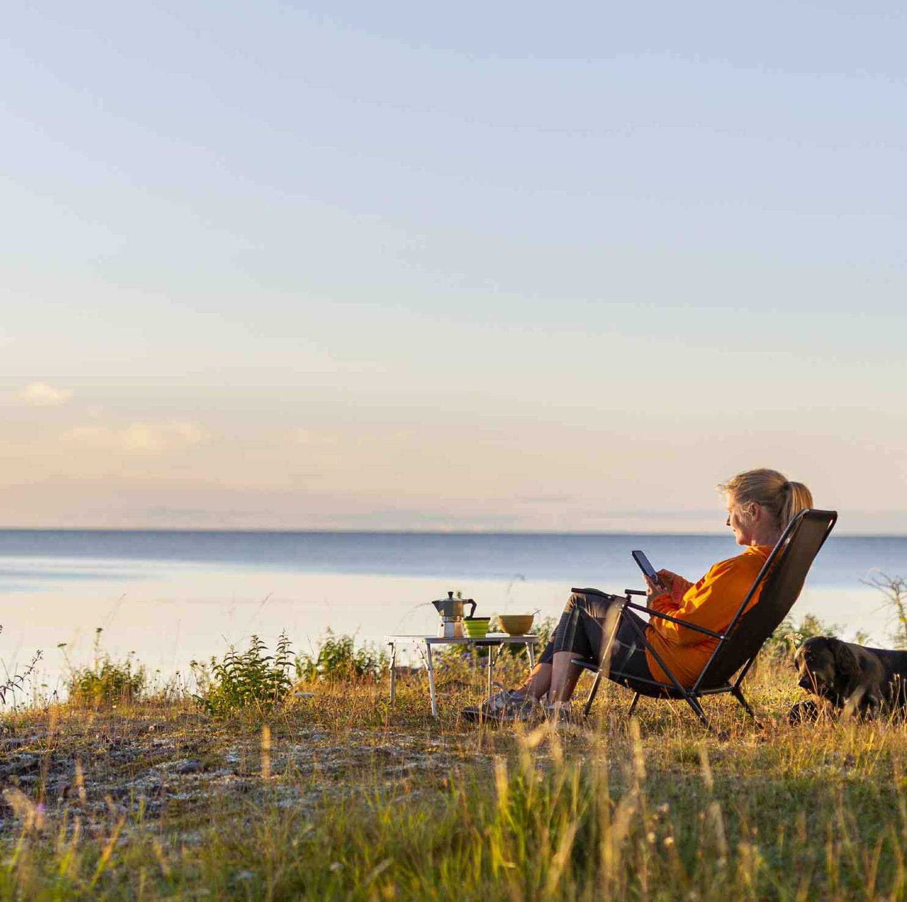 En kvinna i orange jacka sitter i en stol vid en sjö och läder en bok