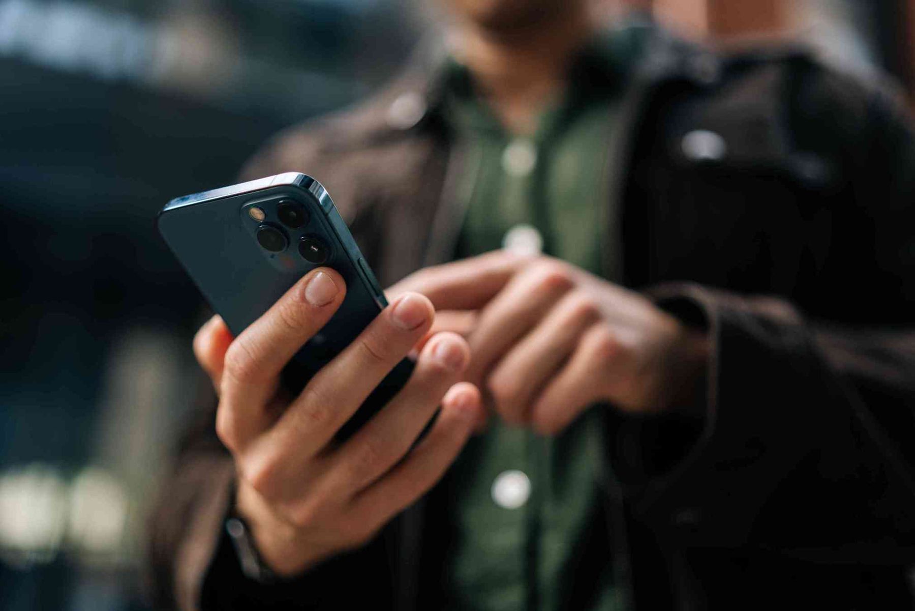 Person använder en smartphone utomhus, klädd i mörk jacka och grön skjorta.