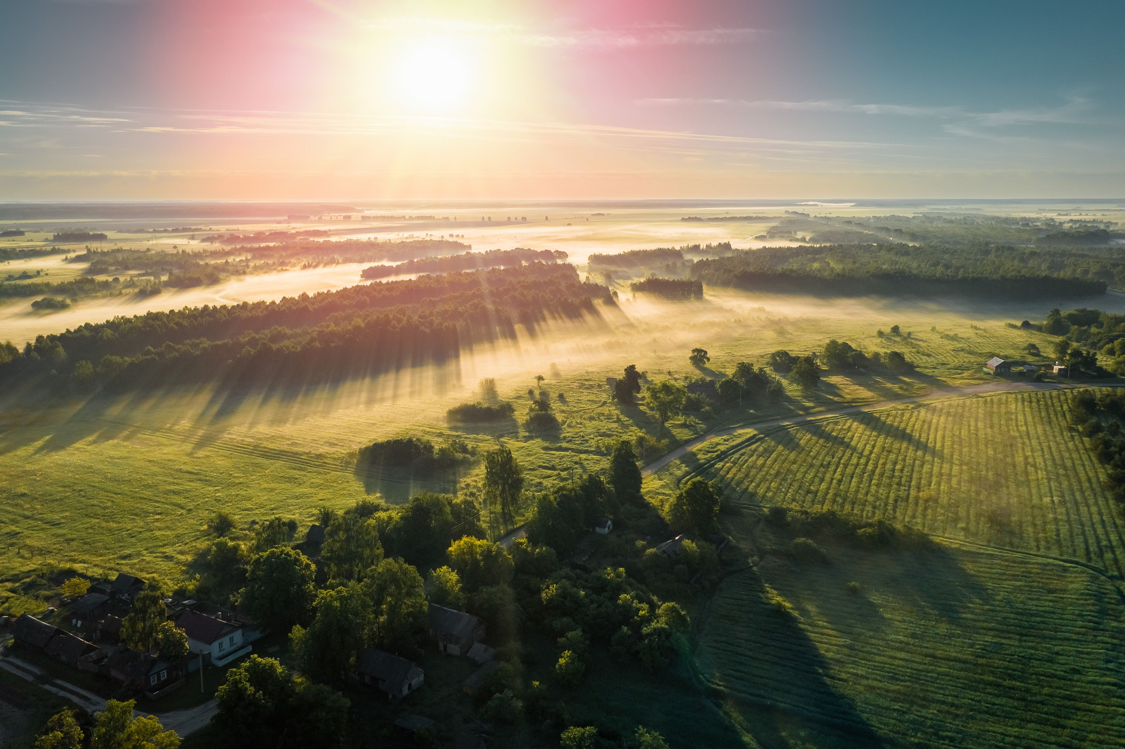 Solskin lyser over et landskab med enge og skove, tågede marker og små huse, hvilket skaber lange skygger og en stemningsfuld atmosfære.