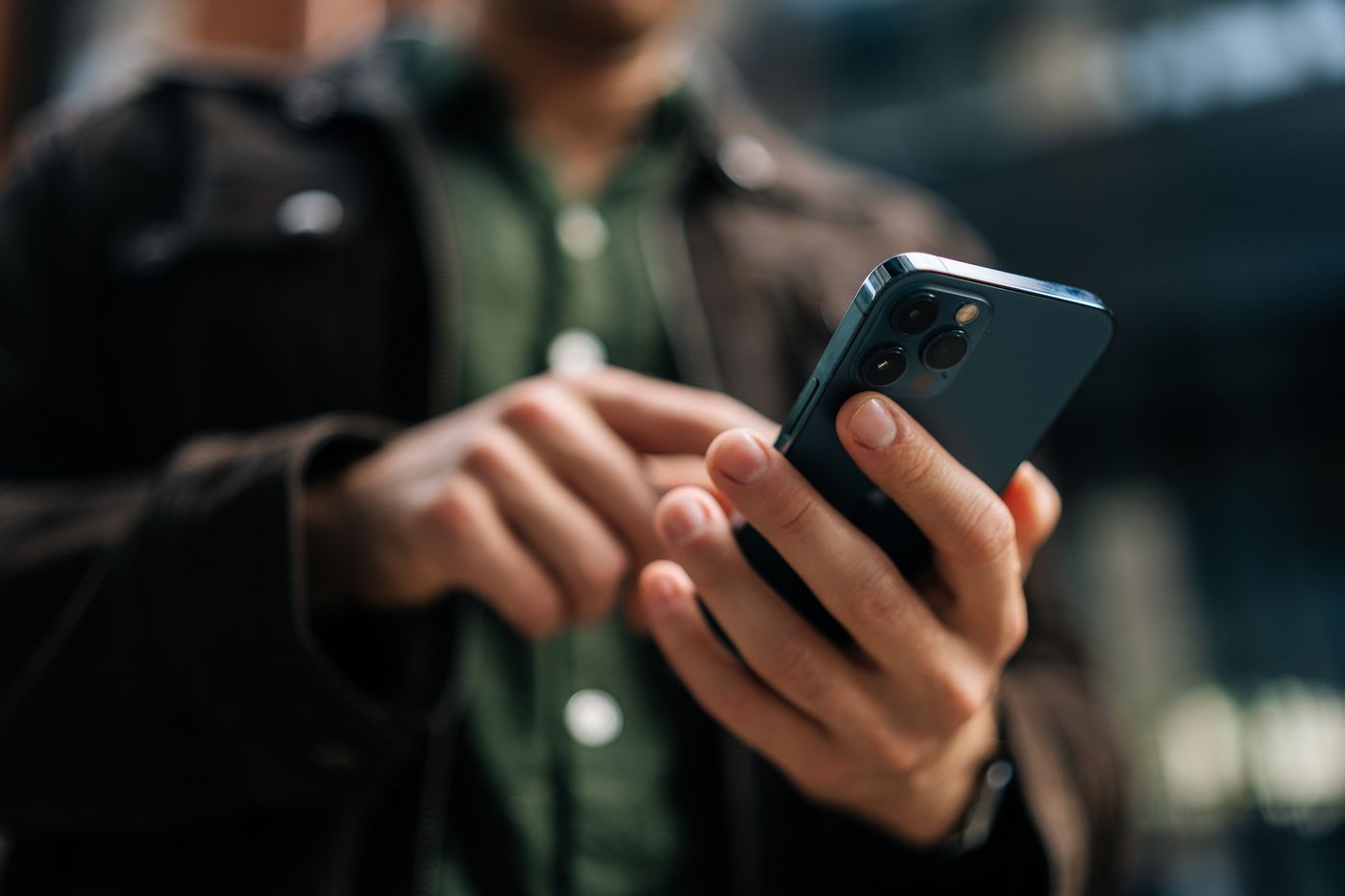 Person håller i en smartphone och använder pekskärmen utomhus, iklädd brun jacka och grön skjorta.
