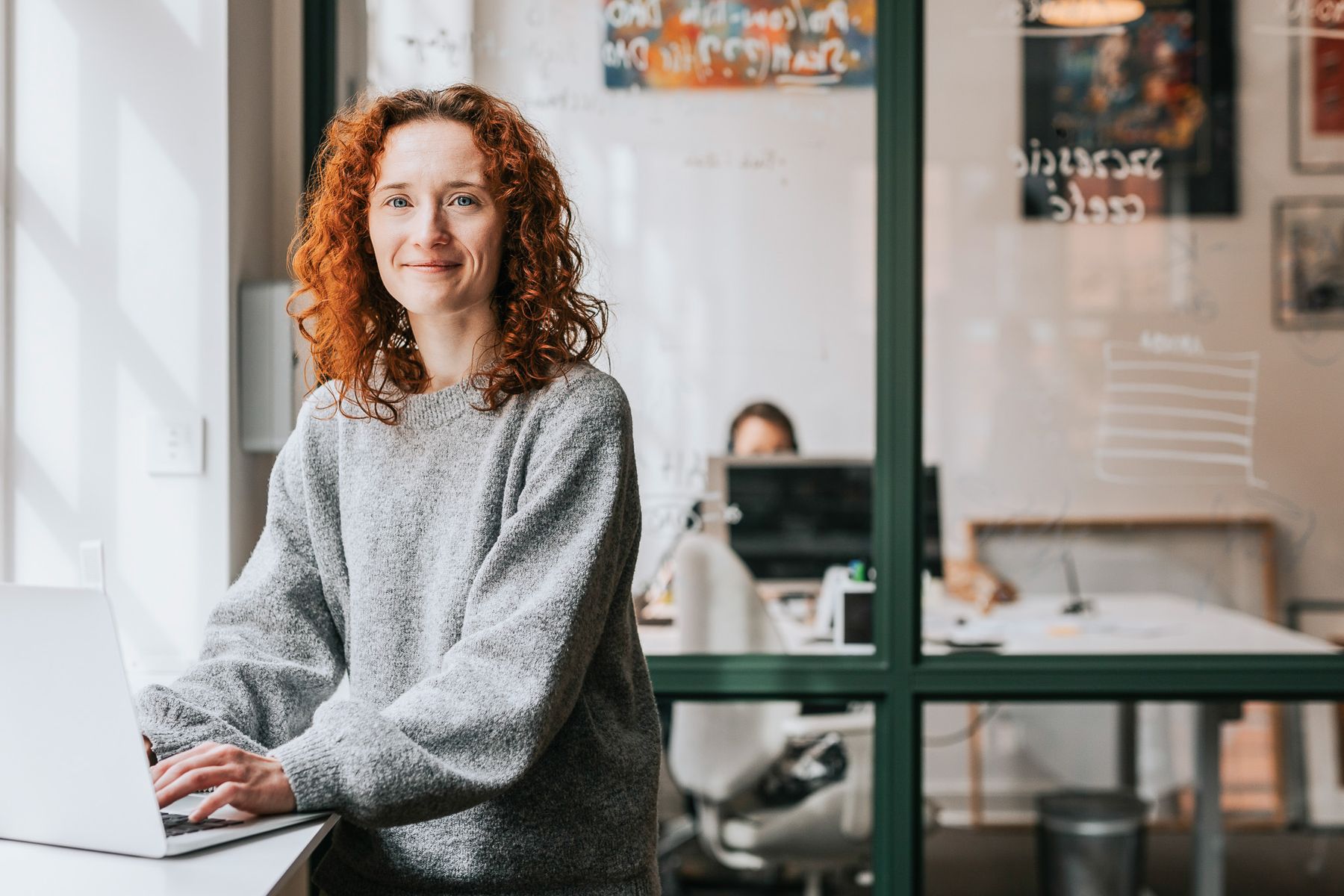 Kvinde med rødt krøllet hår arbejder ved en laptop. Lyse kontormiljøer med glasvægge og en person i baggrunden.