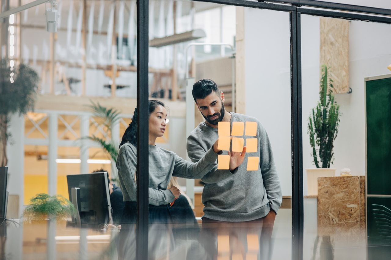 Two people in an office discuss notes on a glass wall, surrounded by computers and plants.