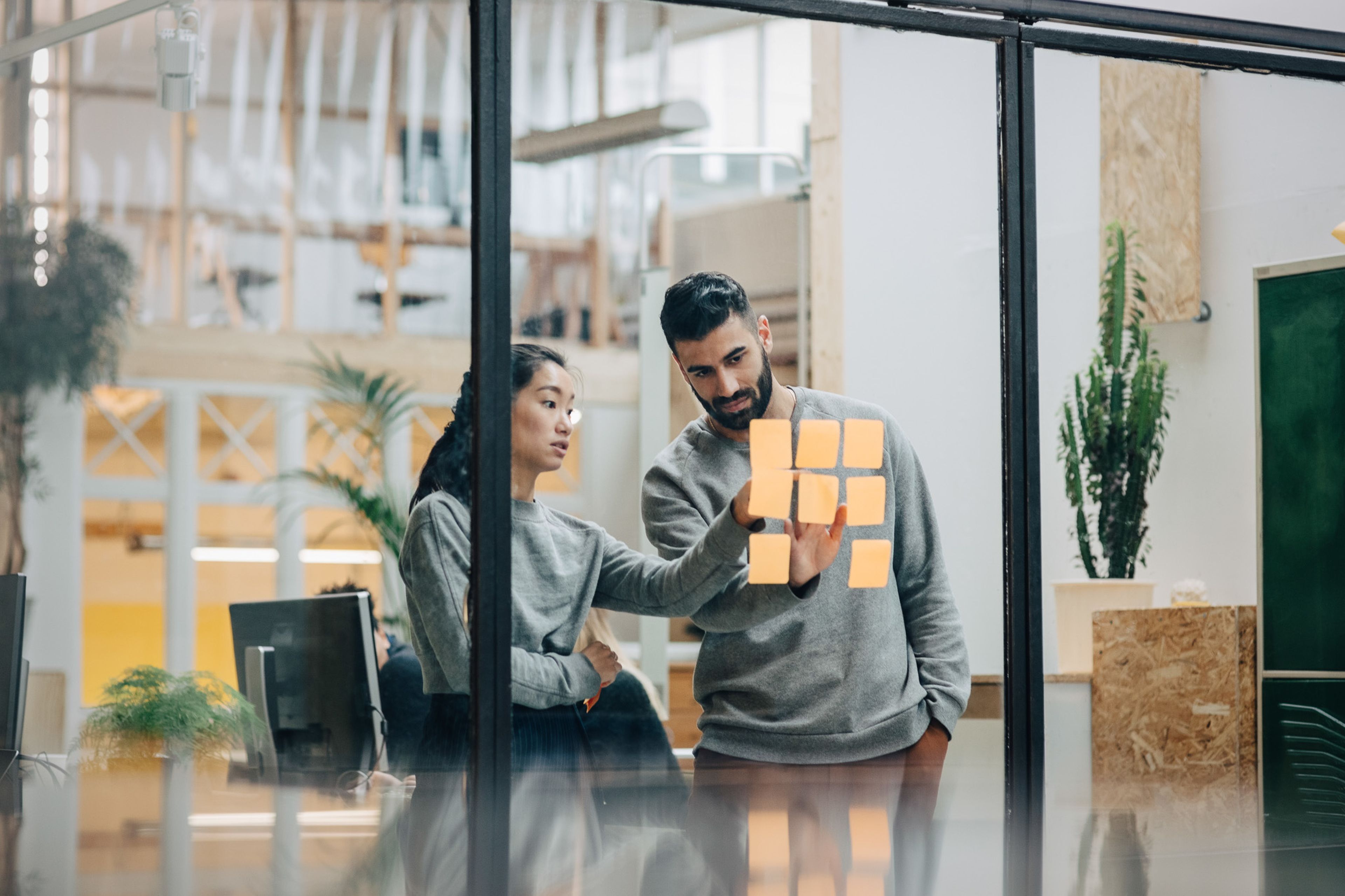 Two people in an office discuss notes on a glass wall, surrounded by computers and plants.
