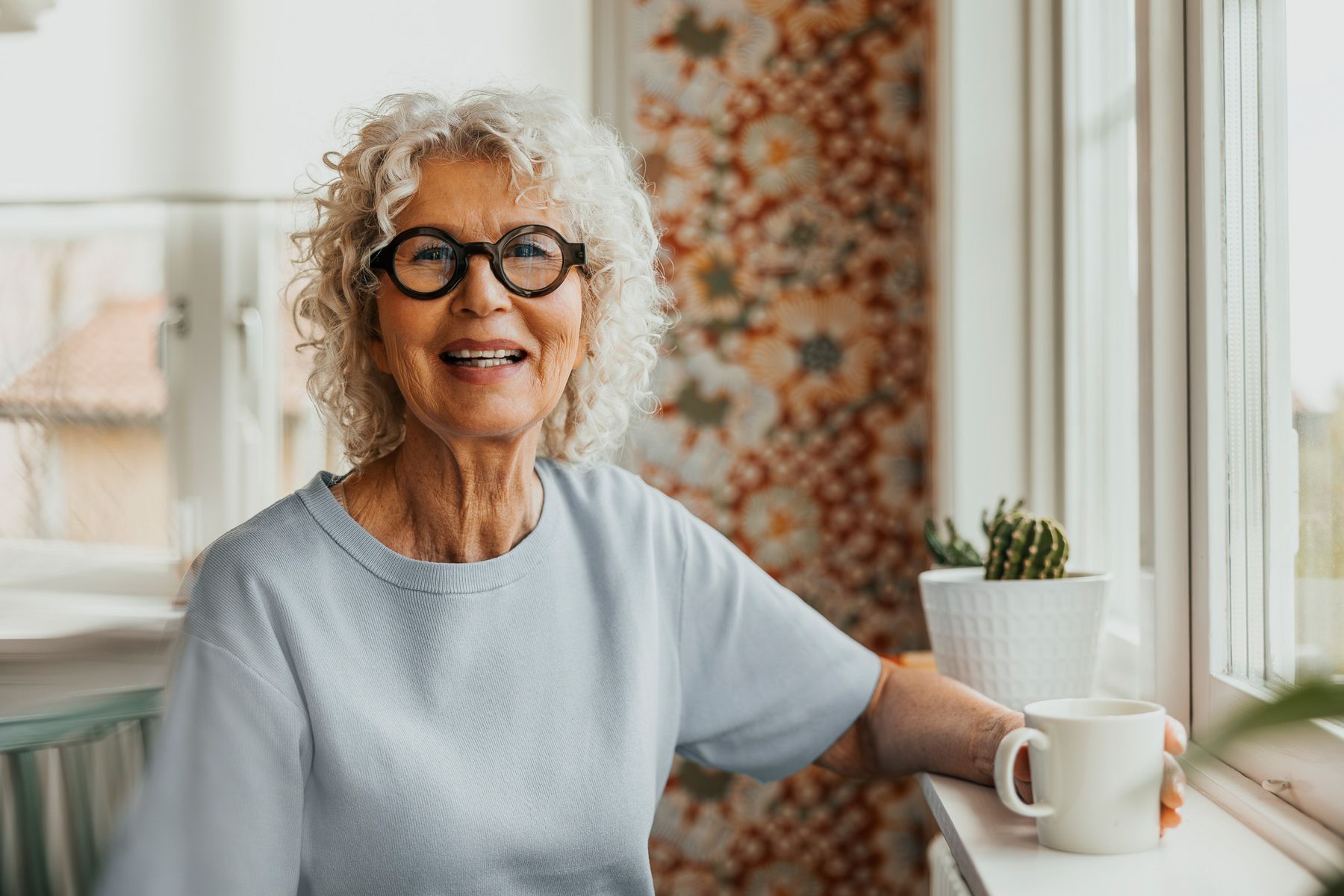 En eldre kvinne med hvitt, krøllete hår og runde briller smiler varmt mot kameraet. Hun lener seg mot en vinduskarm og holder en hvit kaffekrus, med fargerike gardiner i bakgrunnen.