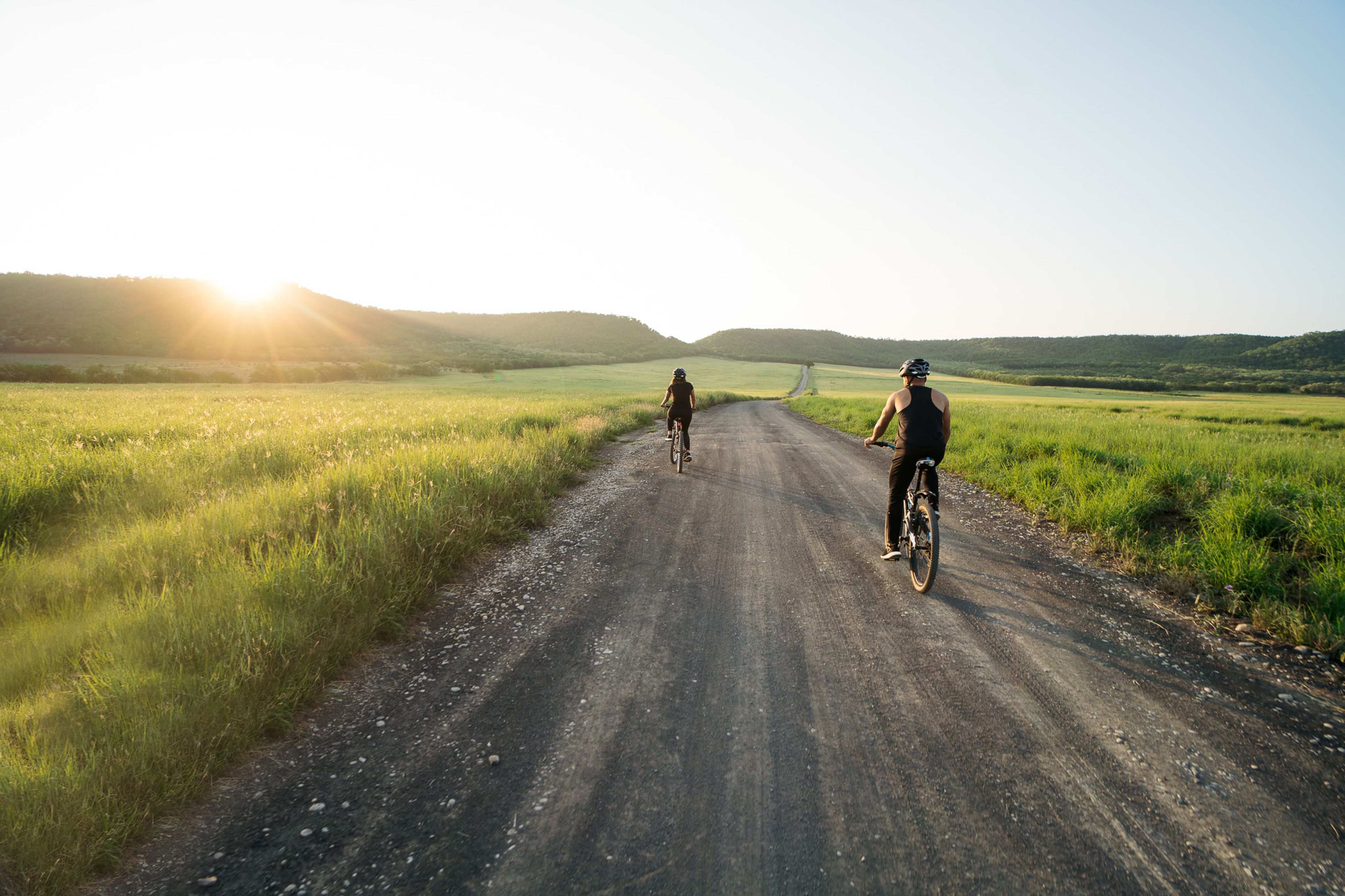 Två cyklister på en landsväg i solnedgången, omgivna av grönska och kullar i bakgrunden.