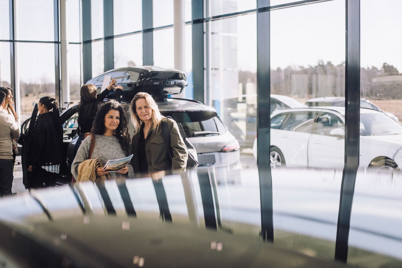 Kaksi naista seisoo autoliikkeessä ja katsoo ajoneuvoja, ympärillään muita ihmisiä ja useita kiiltäviä autoja taustalla.