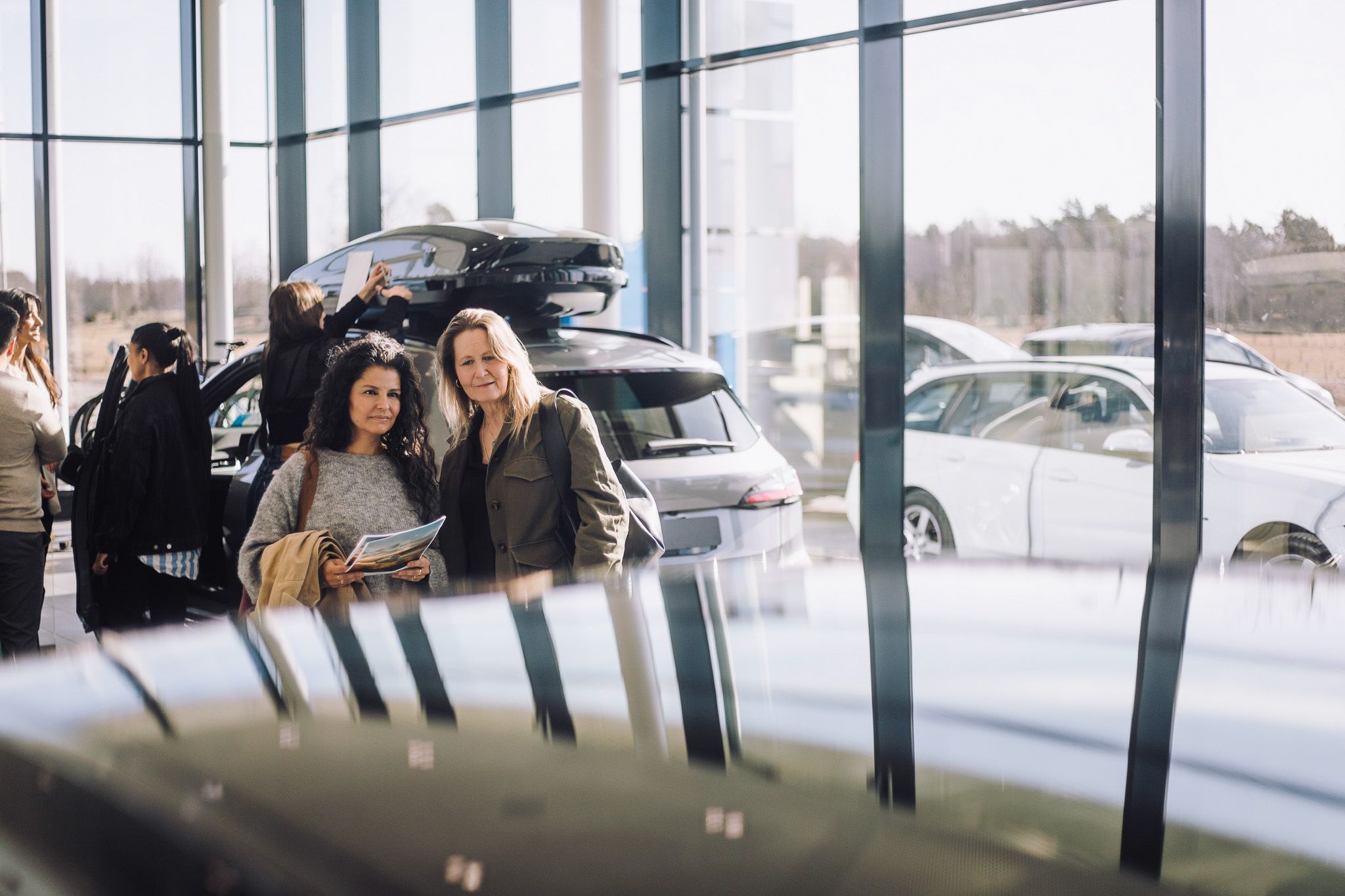 Kaksi naista seisoo autoliikkeessä ja katsoo ajoneuvoja, ympärillään muita ihmisiä ja useita kiiltäviä autoja taustalla.