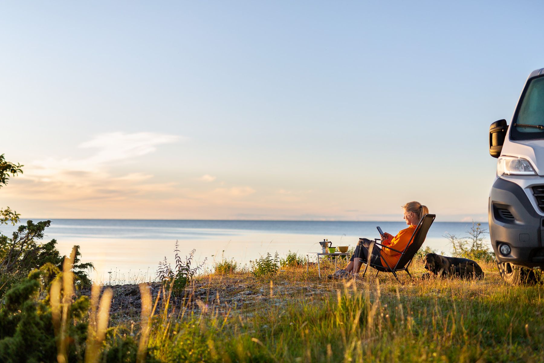 Kvinna sitter i solstol vid havet, använder mobil. En bil och en hund syns bredvid. Gräs och hav i bakgrunden.