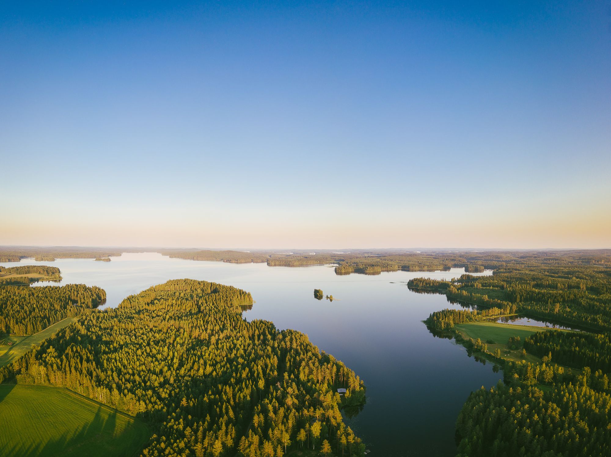 Flygfoto av ett stort landskap med sjöar och öar, omgivna av tät skog och klar blå himmel i soluppgång.