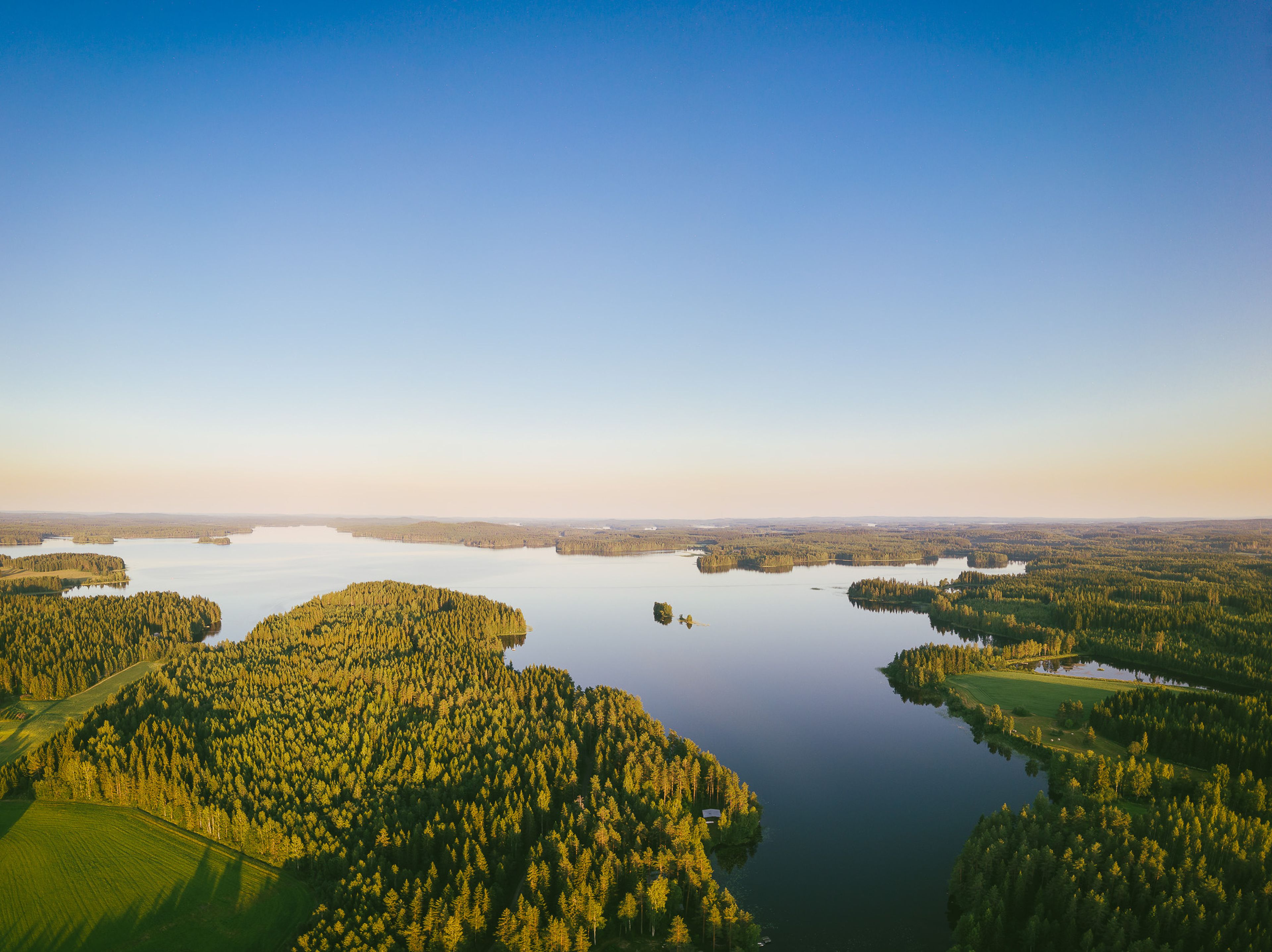 Flygfoto av ett stort landskap med sjöar och öar, omgivna av tät skog och klar blå himmel i soluppgång.