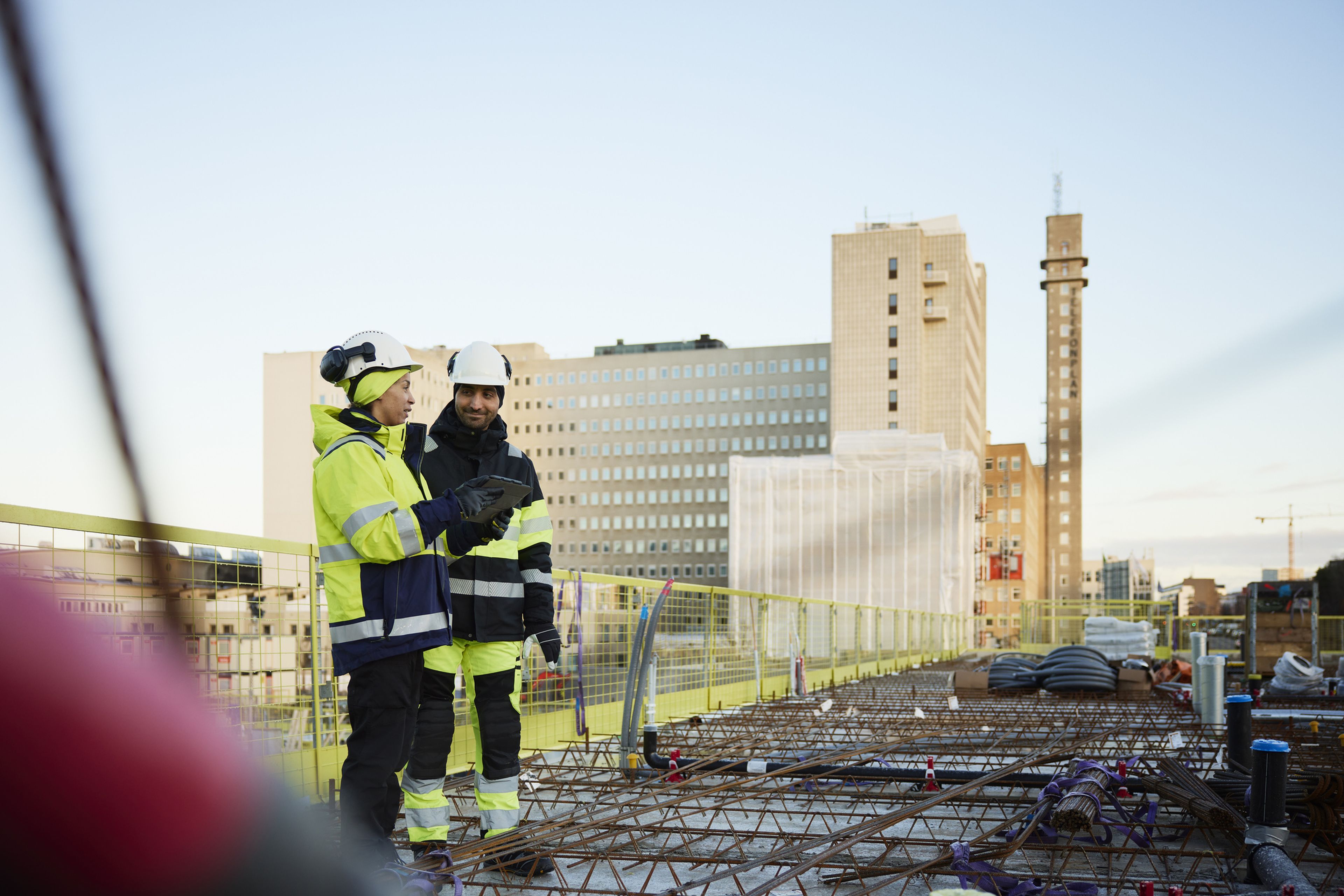 Två byggarbetare i reflexkläder och hjälmar står på en byggplats och diskuterar något på en surfplatta, mot en stadsbakgrund.