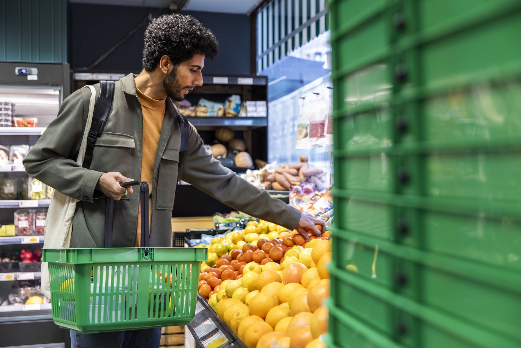 Mann plukker frukt i butikk, holder en grønn kurv. Fruktavdeling med appelsiner og tomater i forgrunnen.