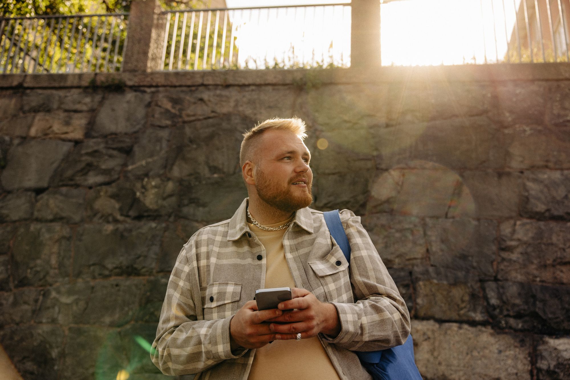 En ung mann med lyst hår og rutet skjorte står utendørs og holder en mobiltelefon. Han ser til siden med et lett smil, og solen skinner kraftig i bakgrunnen bak en steinmur.