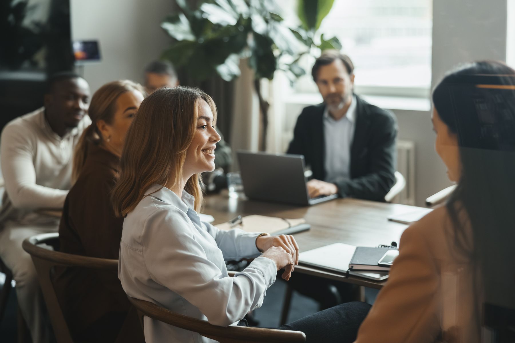 En grupp personer sitter runt ett bord på ett kontor, en kvinna ler och pratar, andra lyssnar uppmärksamt.