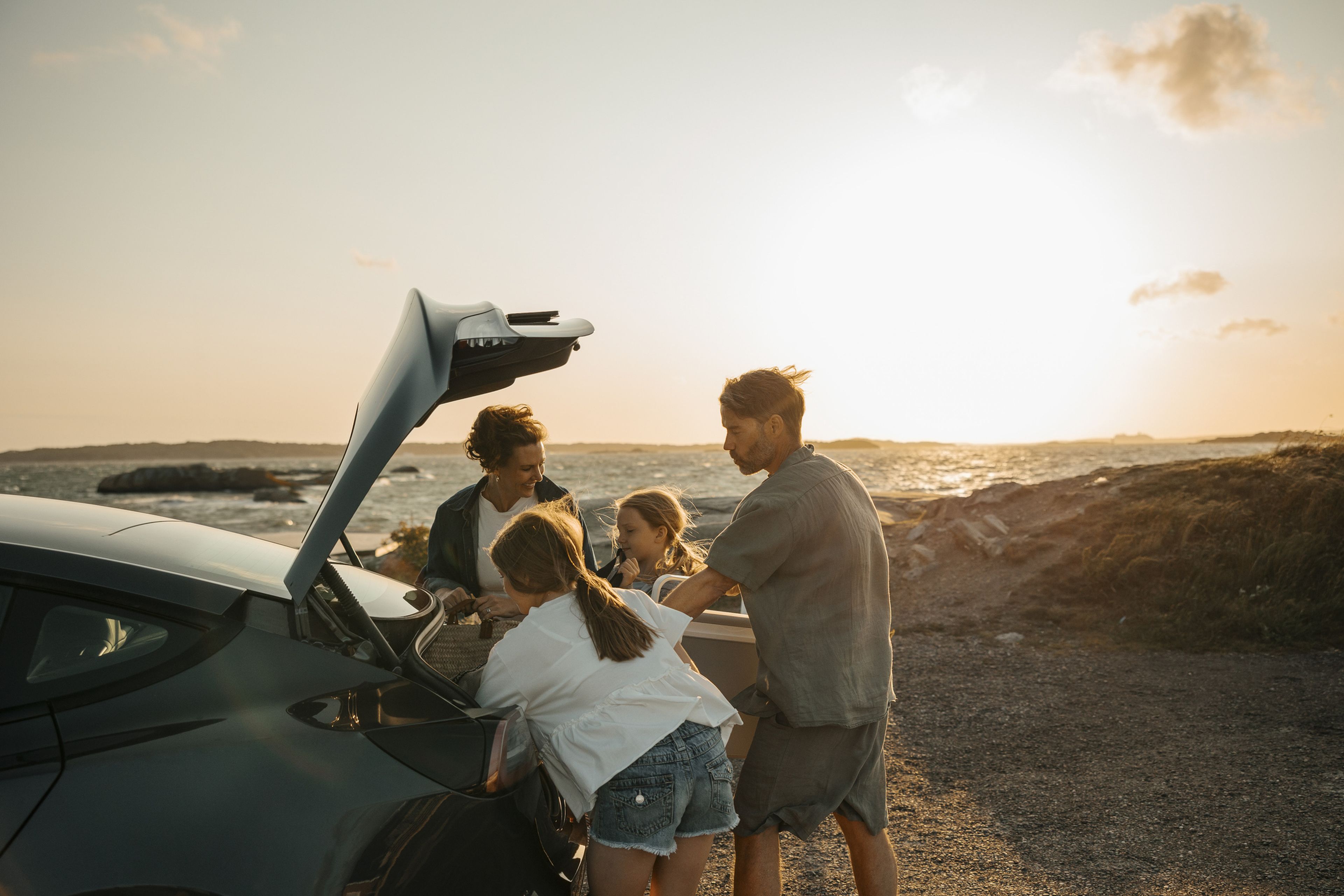 Familj lastar in i modern bil som står parkerad vid havet på västkusten. Det blåser och är solnedgång.