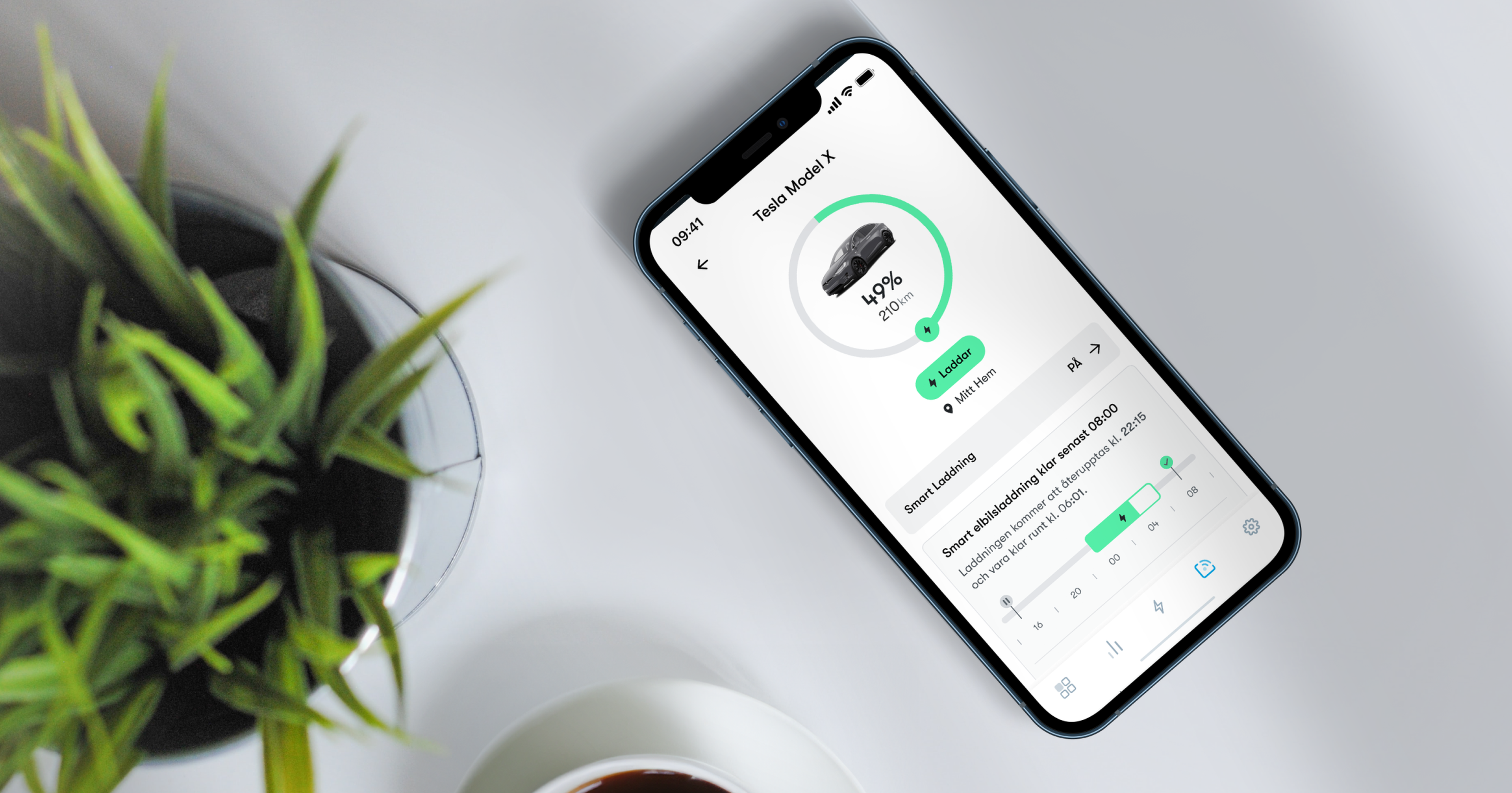 Smartphone visar Tesla Model X-app med 49% batterinivå. Omgiven av en grön växt och en kopp kaffe på ett vitt bord.