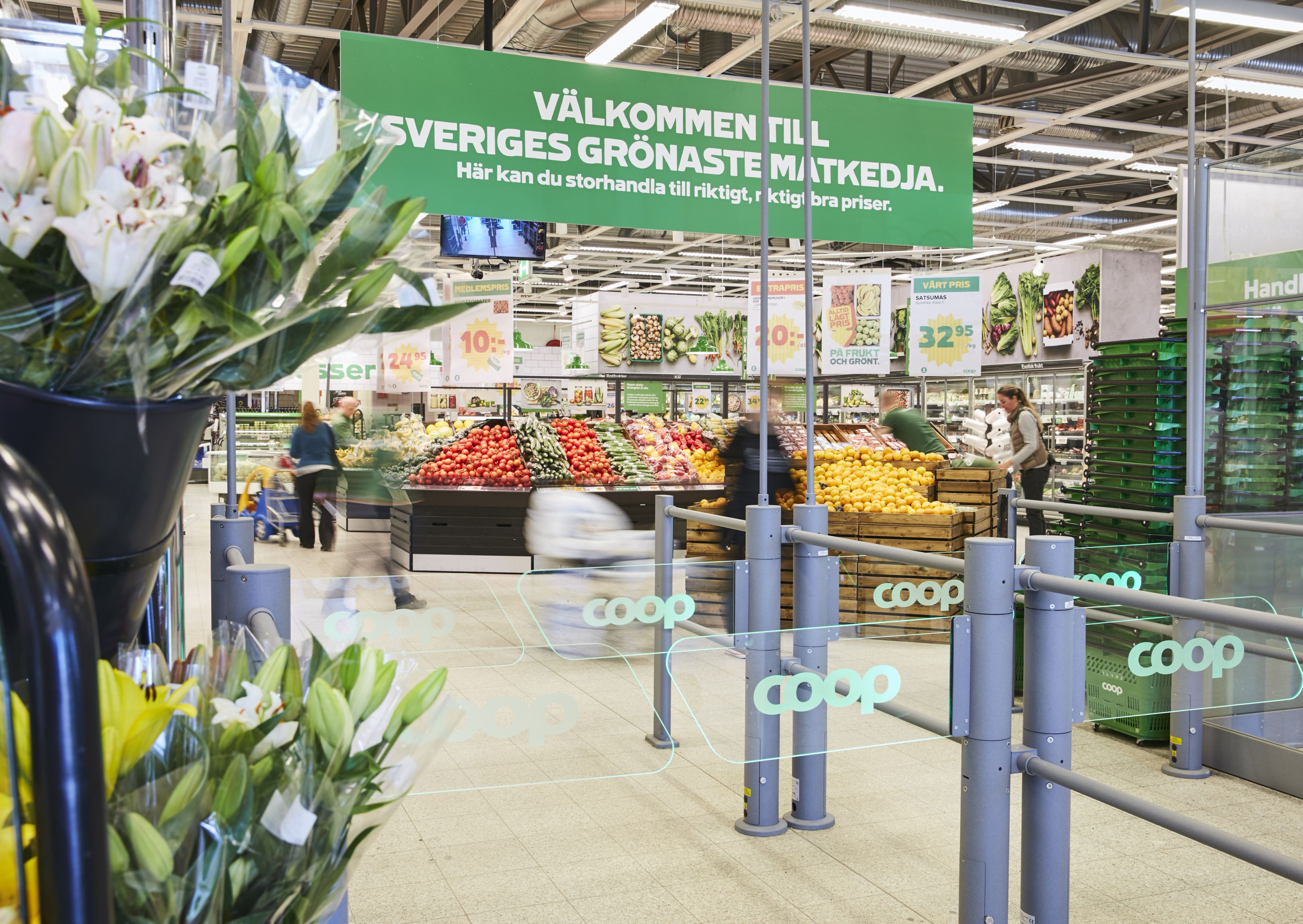 Ingång till Coop med skylt "Välkommen till Sveriges grönaste matkedja". Blommor och fruktavdelning syns i bakgrunden.