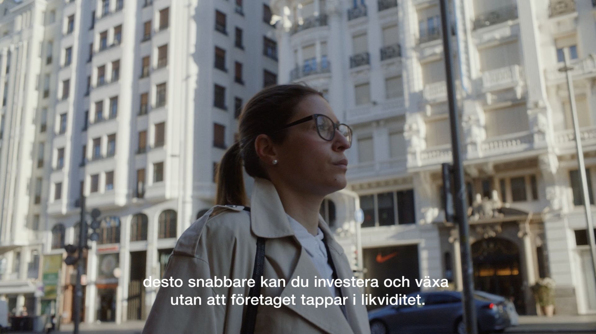 Person i trenchcoat och glasögon går på stadsgata med stora byggnader. Texten säger: "desto snabbare kan du investera och växa utan att företaget tappar i likviditet."