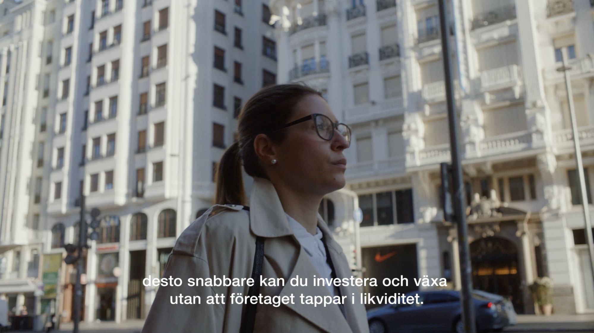 Person i trenchcoat och glasögon går på stadsgata med stora byggnader. Texten säger: "desto snabbare kan du investera och växa utan att företaget tappar i likviditet."