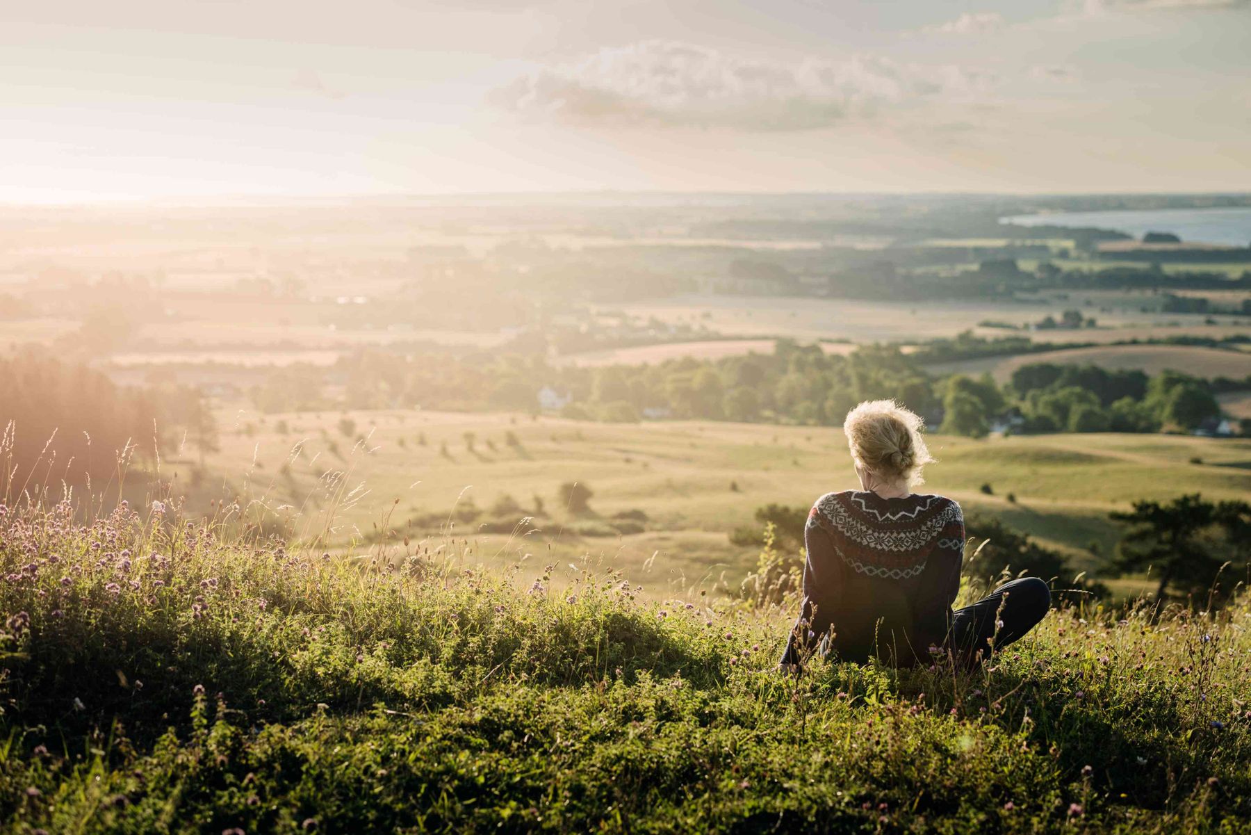 Person sitter på en grønn bakke og ser ut over et vidstrakt landskap i solnedgangsbelysning.