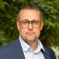 Man med glasögon och skäggstubb, iklädd en kostym och skjorta, mot en bakgrund av gröna träd.