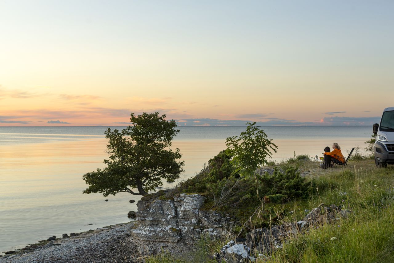 En person i orange jacka sitter i en campingstol vid en stenig kust och blickar ut över ett stilla hav i solnedgångens varma, pastellfärgade ljus. Bredvid syns ett ensamt lövträd och en husbil. I bakgrunden tonar himlen i nyanser av guld, rosa och ljusblått.