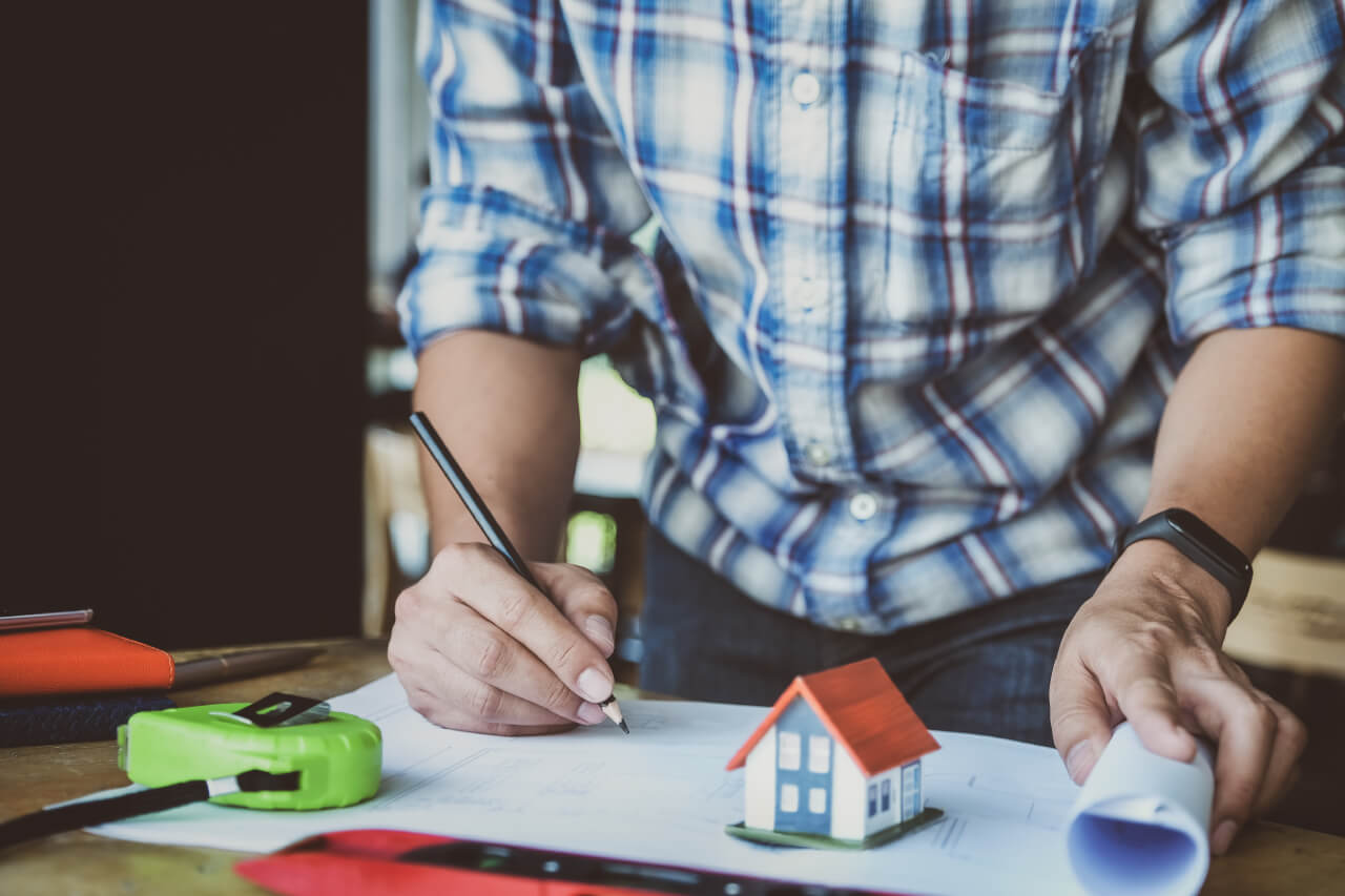 Kockás inges személy ceruzával rajzol építészeti tervekre, egy kis ház makettje és mérőszalag mellett az asztalon.