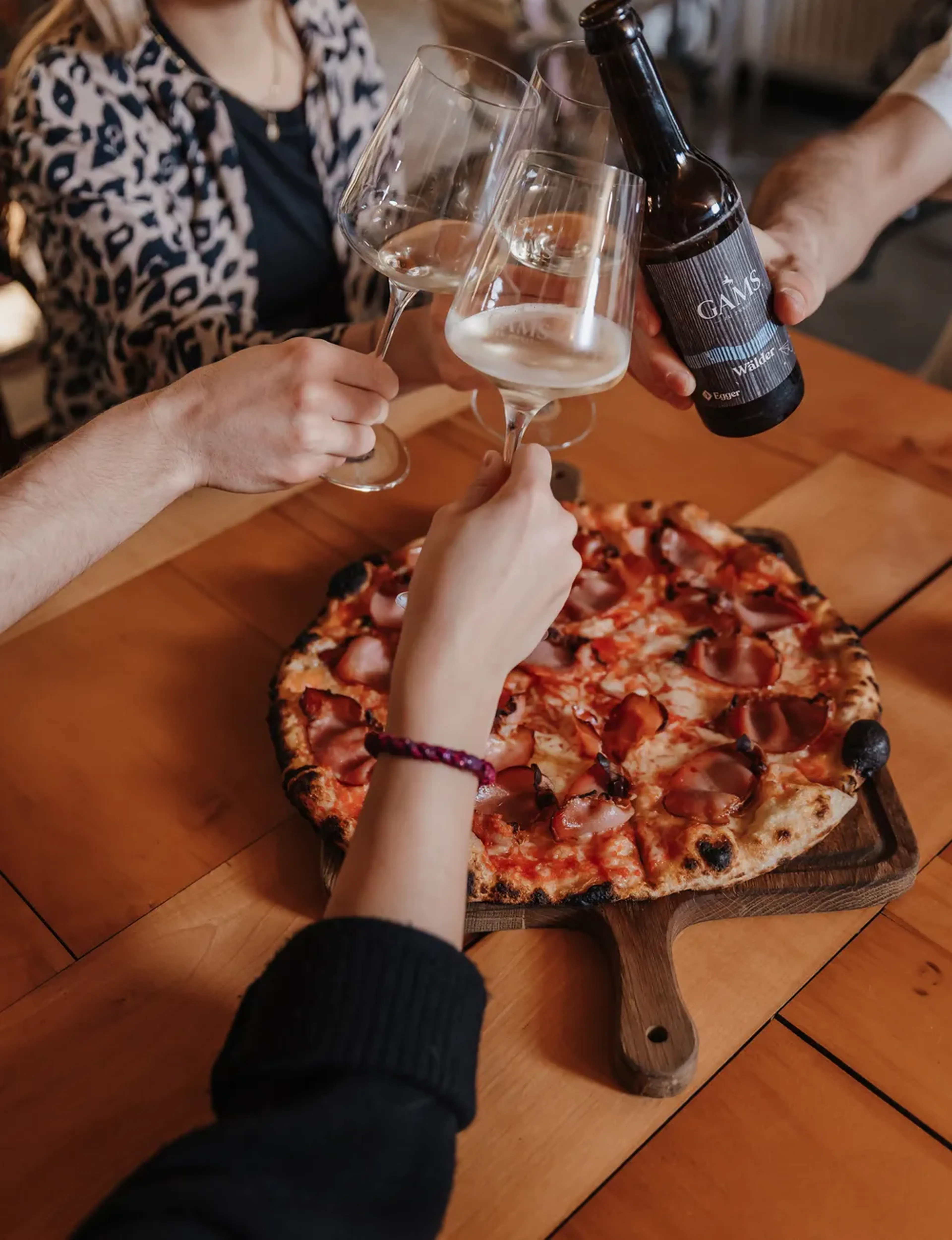 Menschen stoßen mit Weingläsern und einer Bierflasche über einer Pizza auf einem Holztisch an.