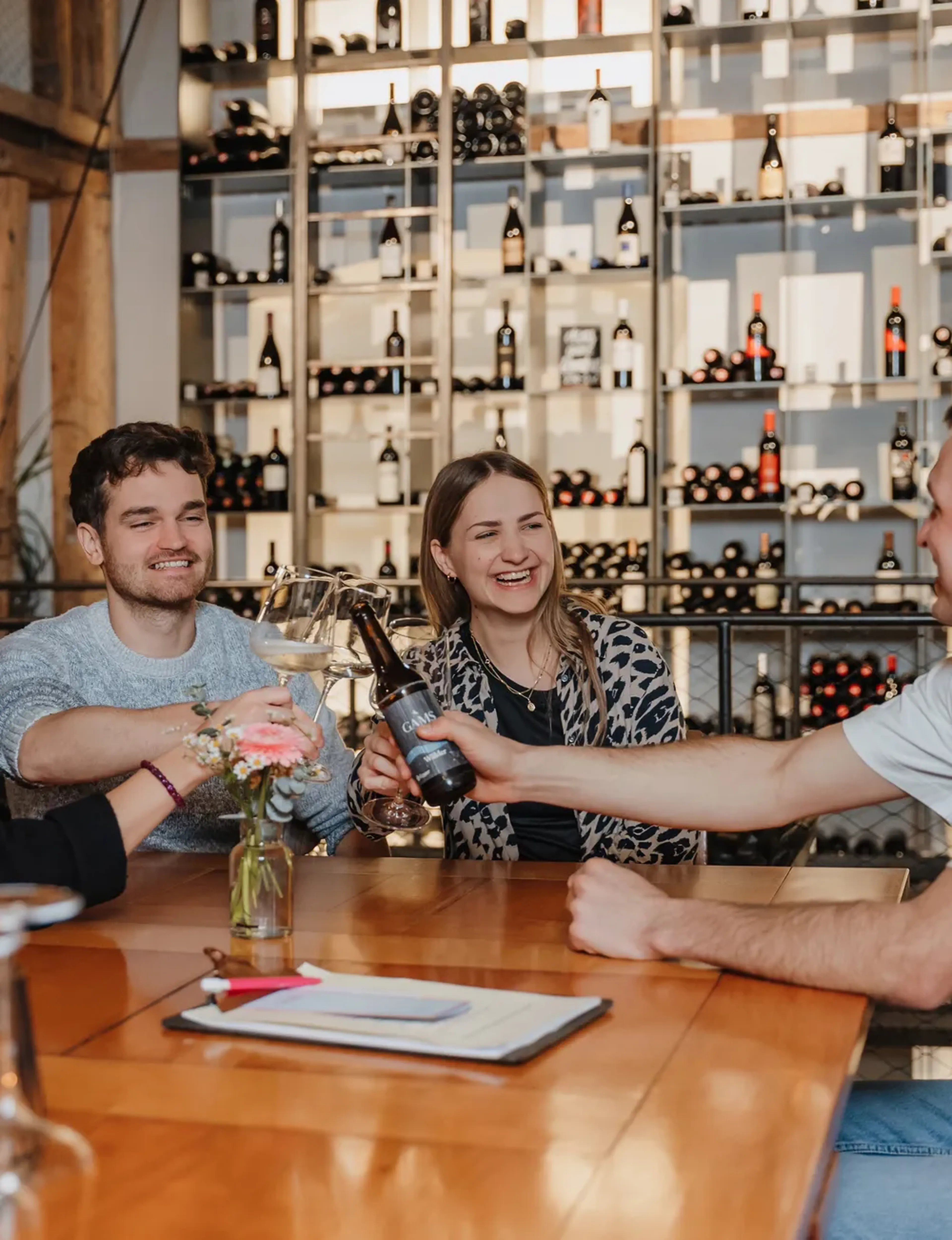 Drei Personen stoßen mit Gläsern und einer Flasche an einem Tisch in einem Weinkeller an. Im Hintergrund sind Weinregale zu sehen.