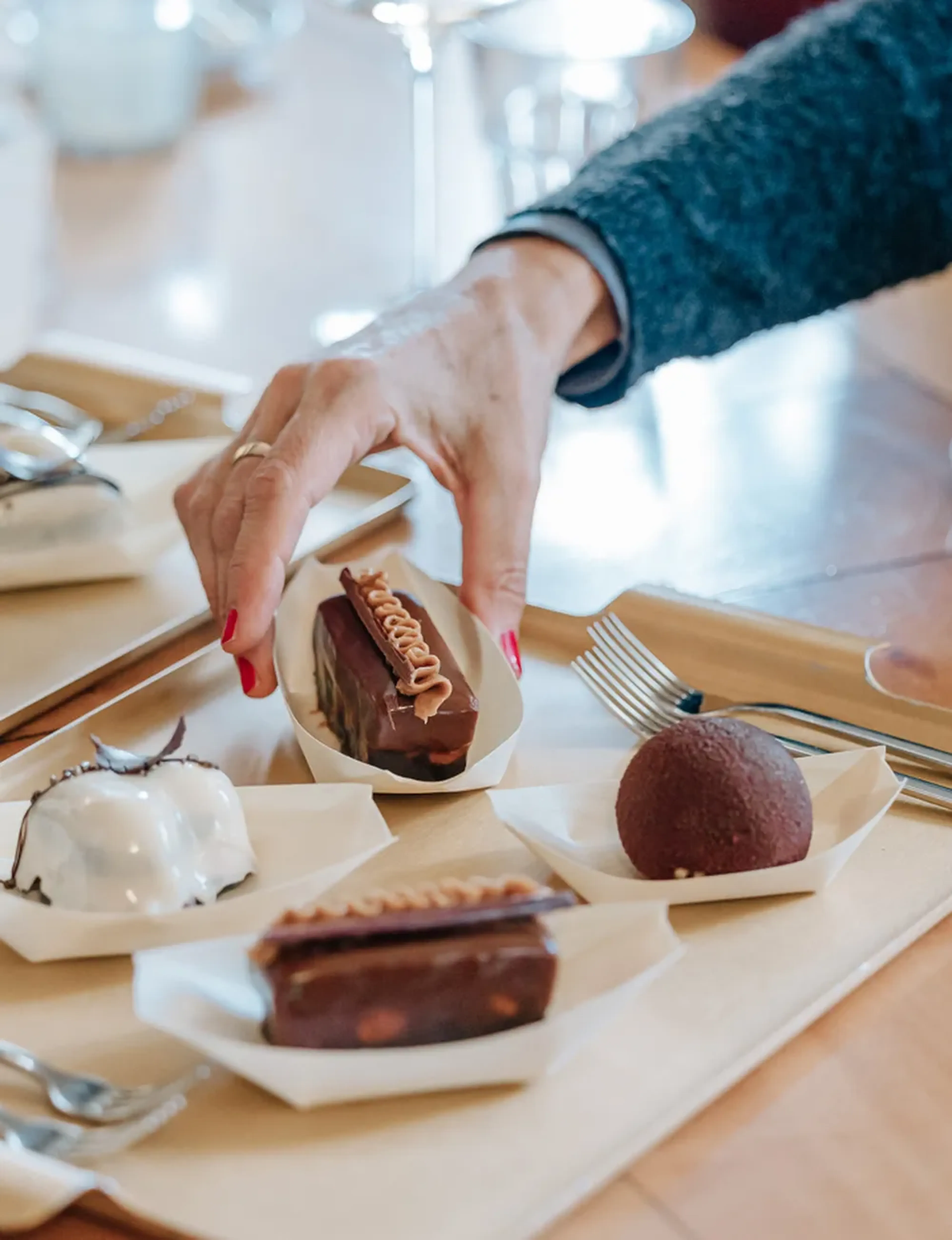 Eine Hand greift nach einem Schokodessert auf einem Tablett mit mehreren kleinen, kunstvoll dekorierten Süßigkeiten.