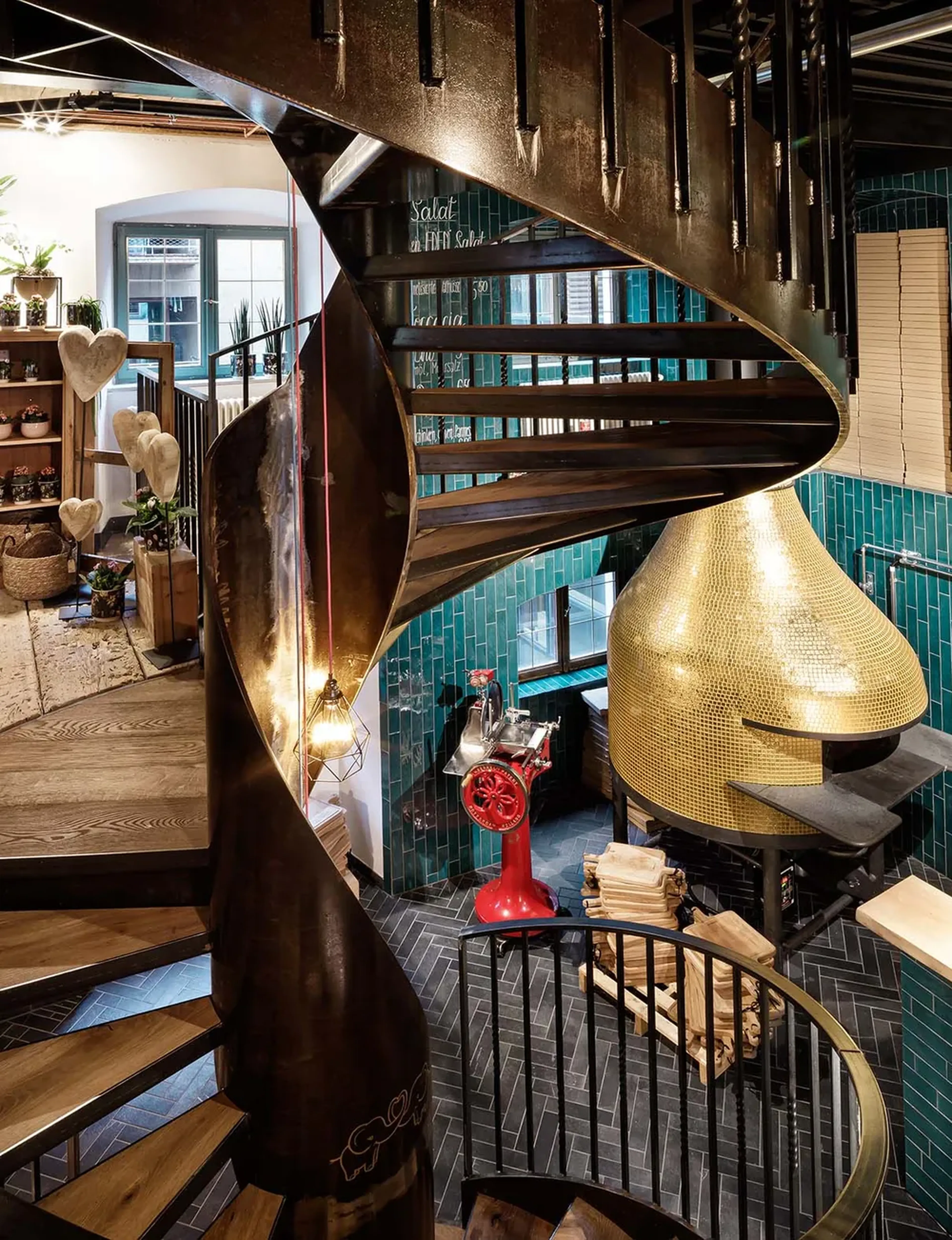 Gewundene Metalltreppe in einem stylischen Raum mit türkisen Fliesen, einem roten Fleischschneider, Holzauslagen und einem goldenen Kuppelofen.