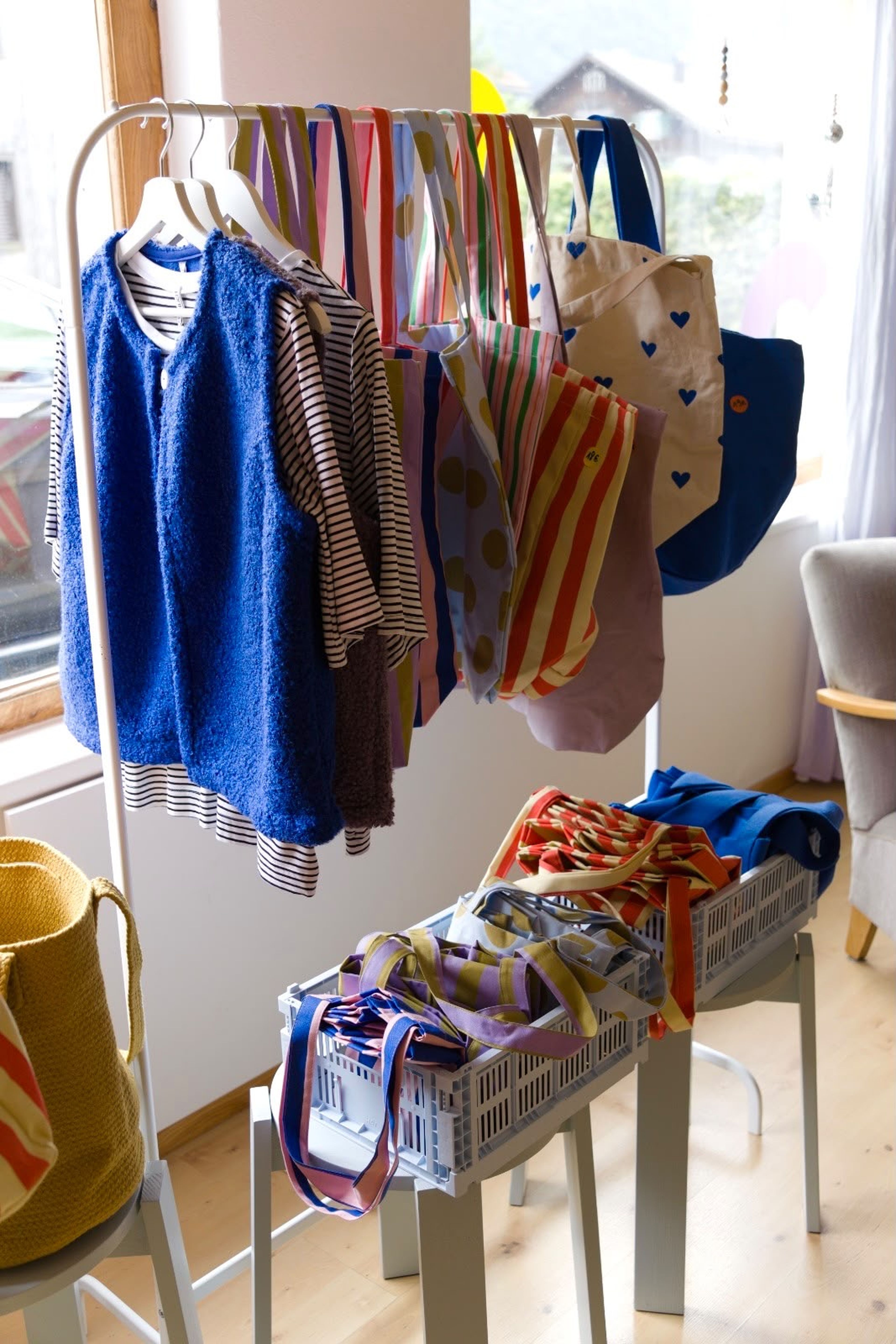 Kleiderständer mit gestreiften Hemden und blauer Weste. Daneben Kisten mit bunten gestreiften Stofftaschen in einem hellen Raum.