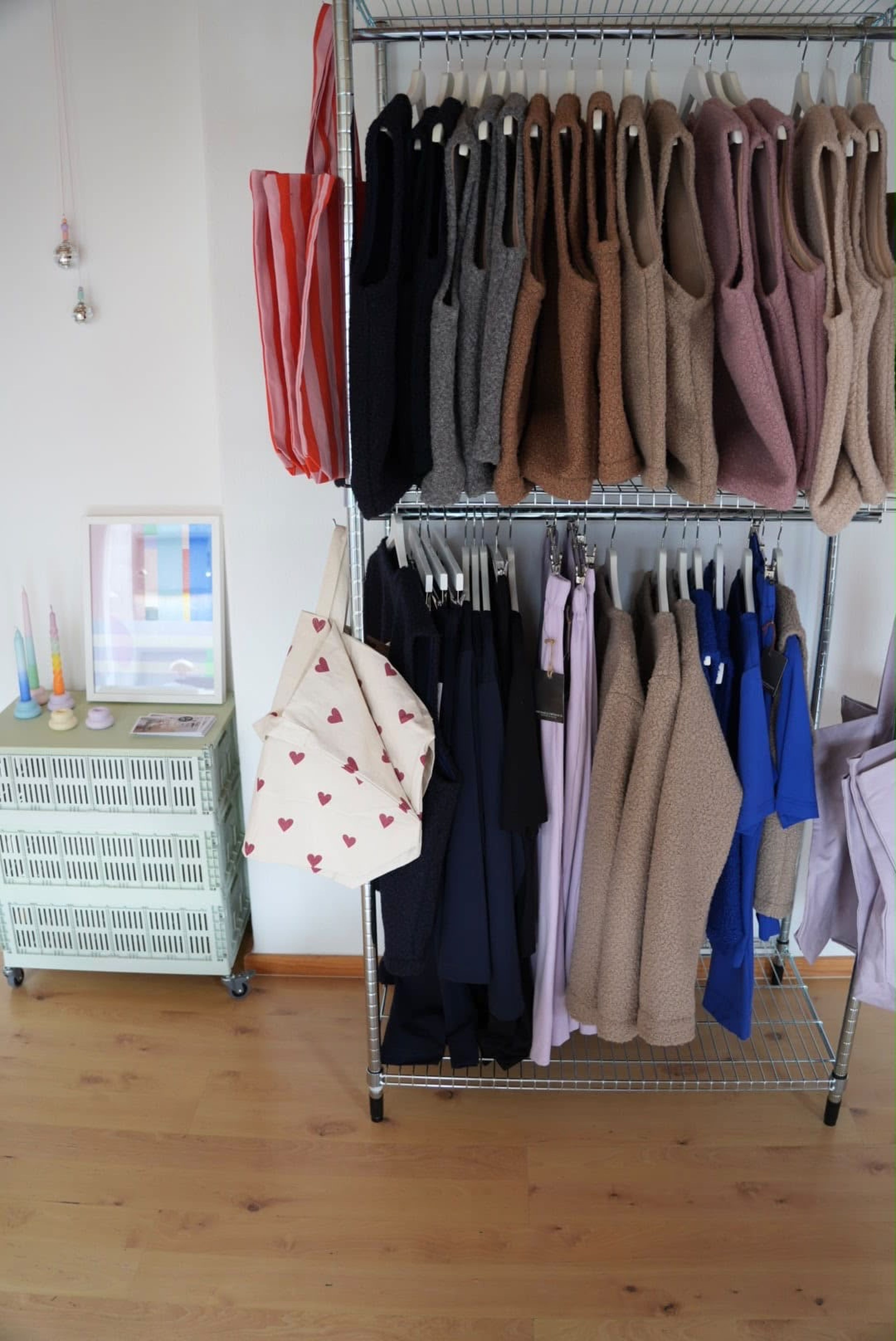 Kleiderständer mit verschiedenen Oberteilen in neutralen Farben, daneben Taschen; pastellfarbene Dekoration auf mintgrüner Kommode. Holzboden.
