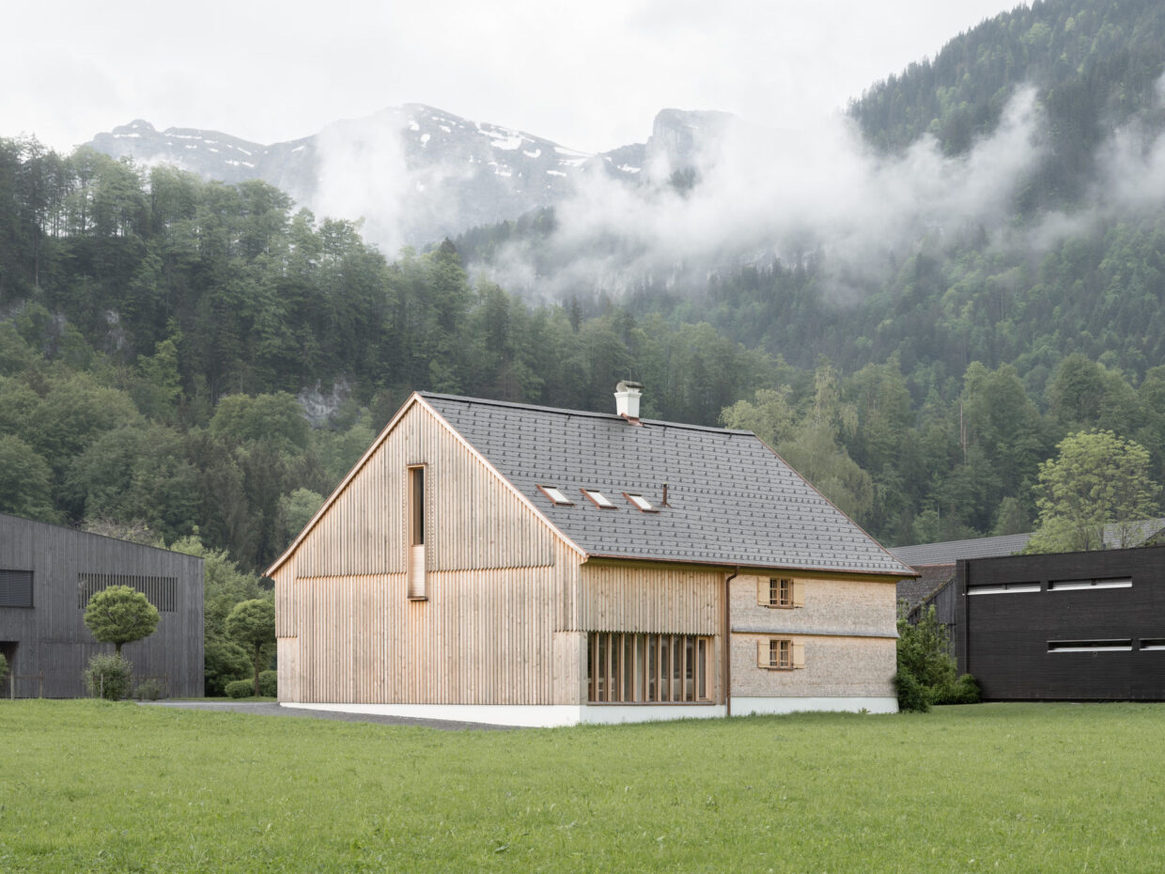 Ein modernes Holzhaus steht auf einer grünen Wiese, umgeben von bewaldeten Bergen mit leichtem Nebel und Wolken am Himmel.