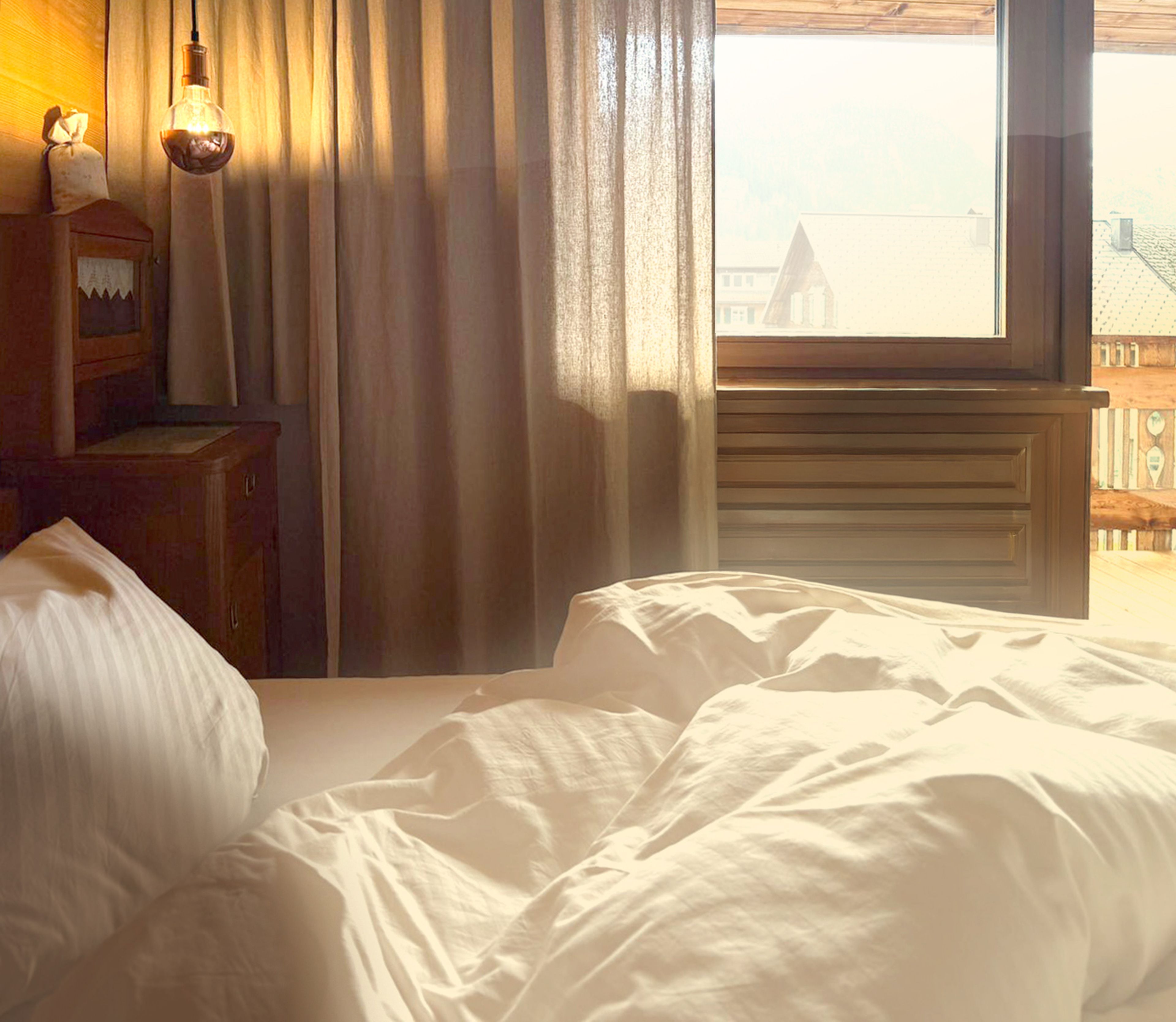 Ein sonnendurchflutetes Schlafzimmer mit ungemachtem Bett, drapierten Vorhängen, einem Nachttisch aus Holz und einem Blick durch das Fenster auf Dächer und Berge.