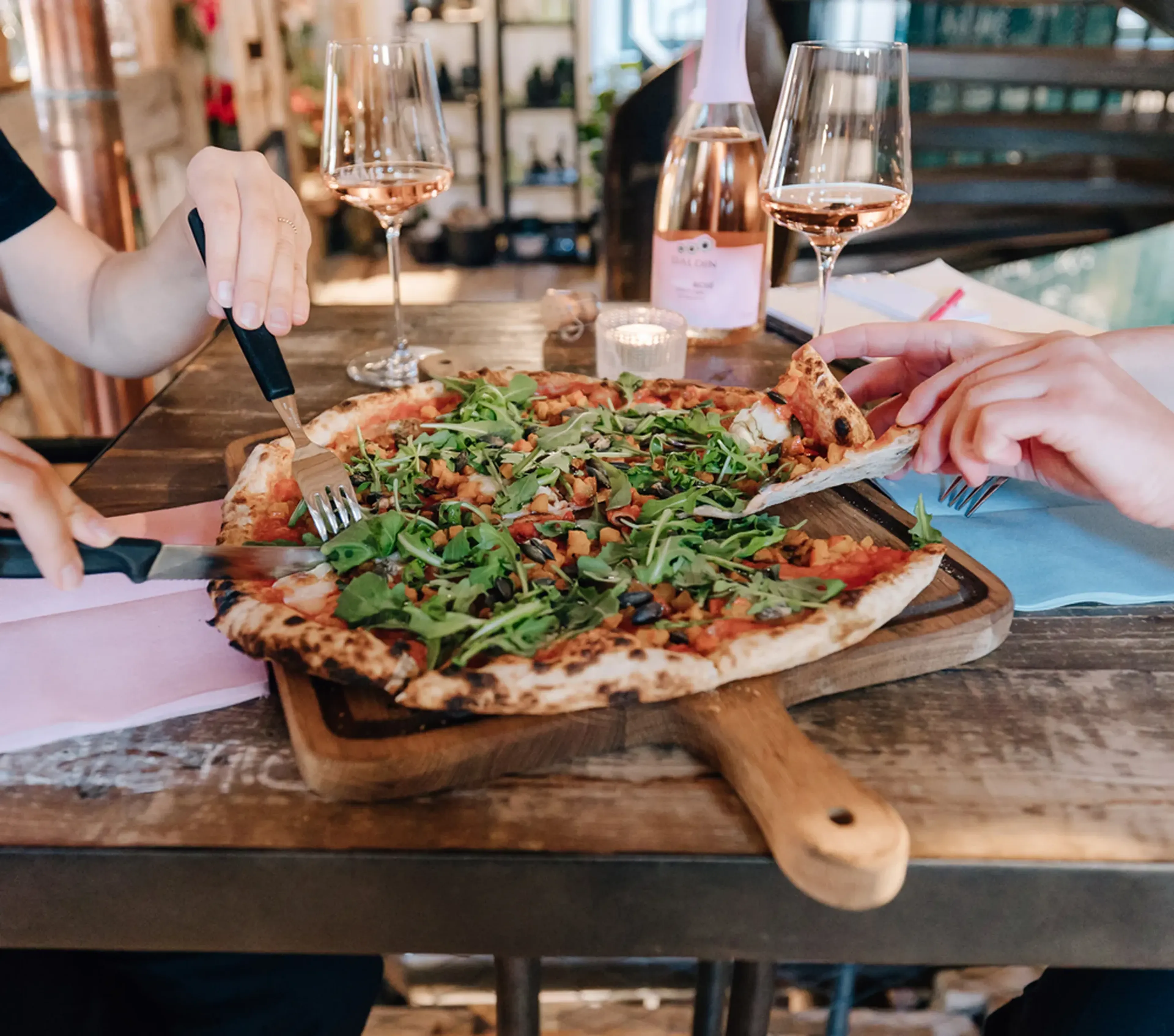 Zwei Personen teilen sich eine Pizza voller Rucola auf einem hölzernen Tablett, mit Roséwein im Hintergrund.