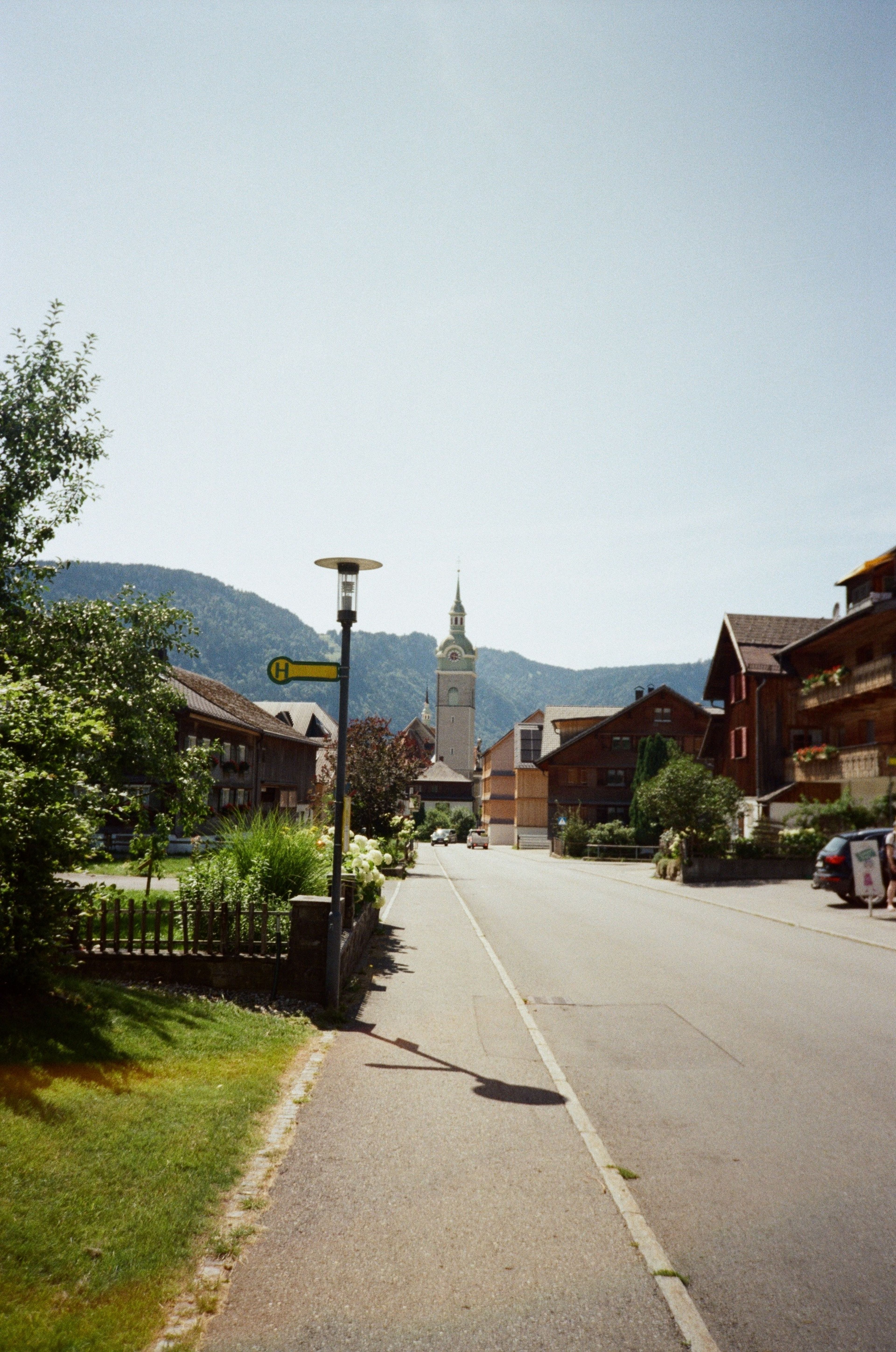 Dorfstraße mit Kirchturm im Hintergrund, umgeben von traditionellen Häusern und Bergen, unter einem klaren, blauen Himmel.