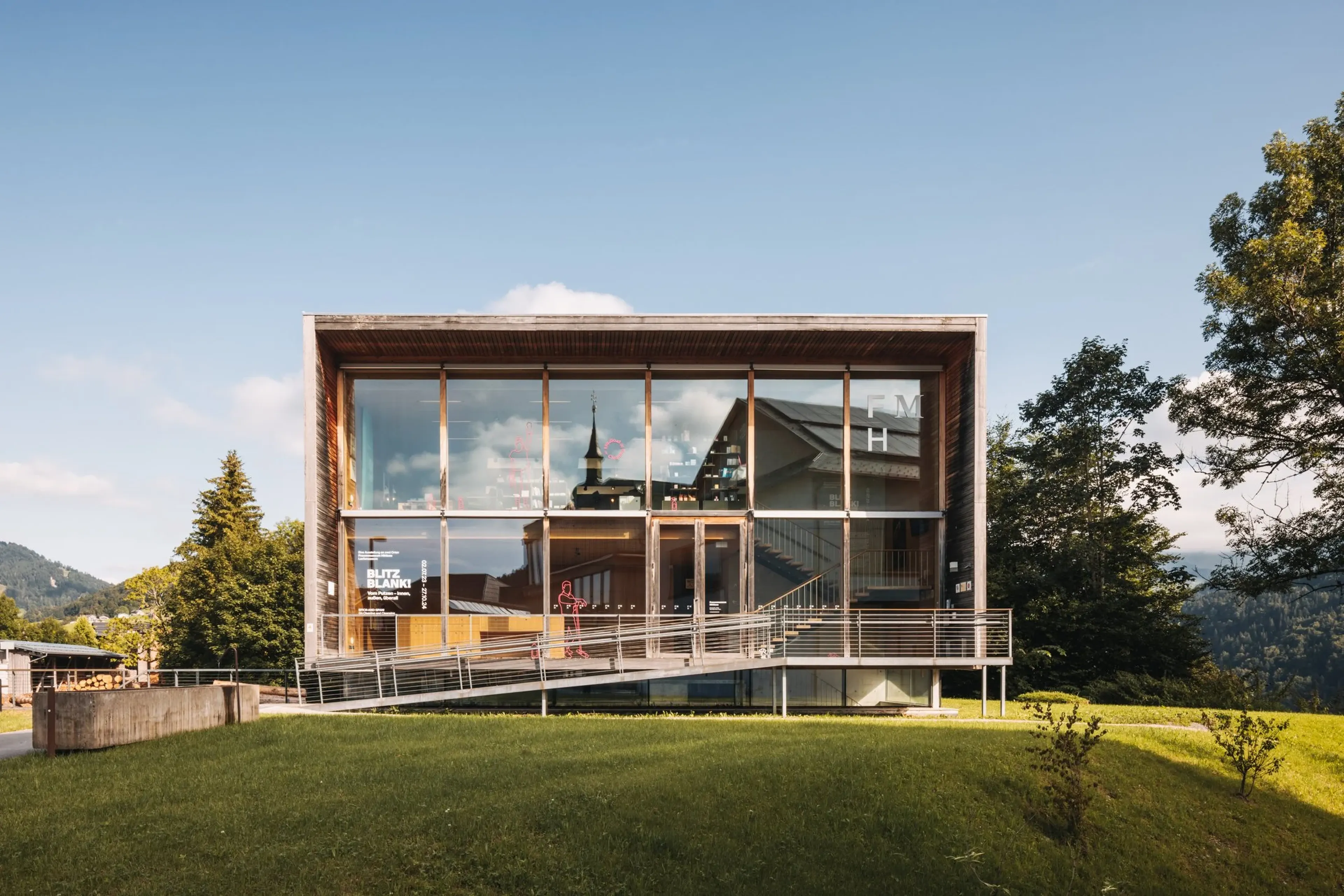 Modernes Gebäude mit Glasfassade und Treppe, eingebettet in eine grüne Landschaft, bei klarem Himmel und Sonnenschein.