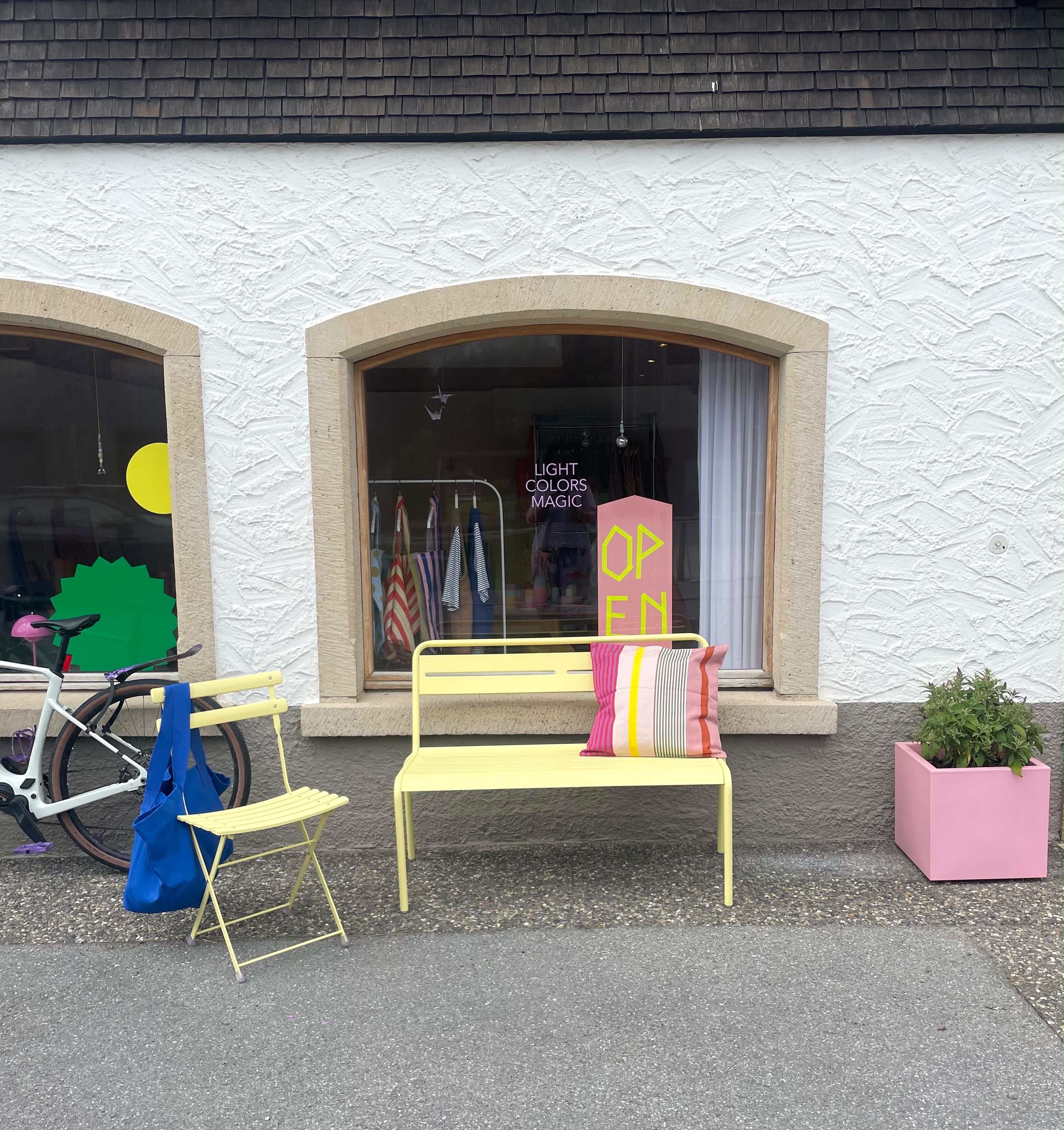 Gelbe Bank und Stuhl vor einem Schaufenster mit Open-Schild. Ein bunt gestreiftes Kissen liegt auf der Bank, daneben ein rosa Blumentopf.